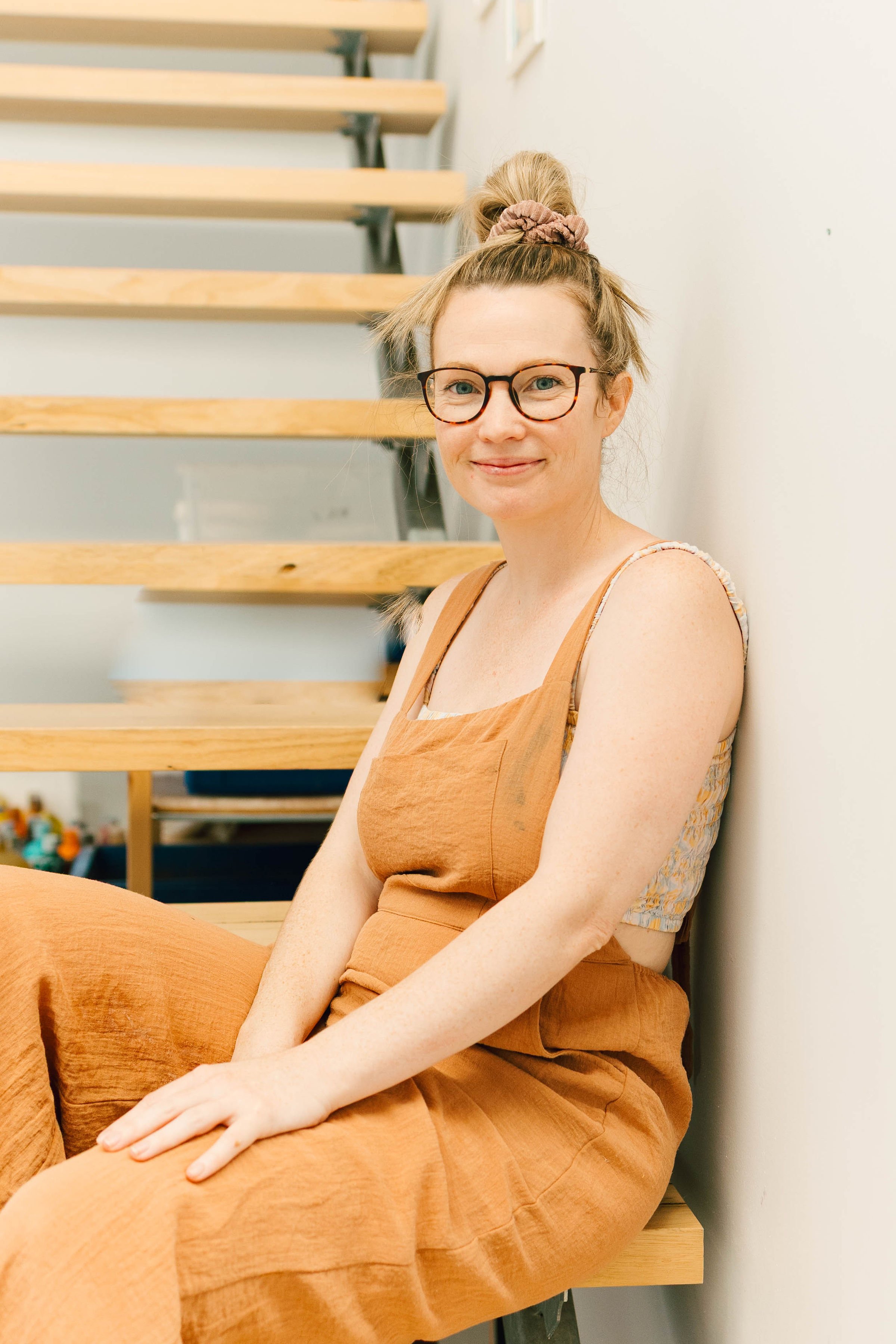 A woman with light skin, glasses, and her hair in a messy bun with a pink scrunchie, sitting on wooden stairs in front of a white wall, smiling at the camera. She is wearing a sleeveless top and a burnt orange dress.
