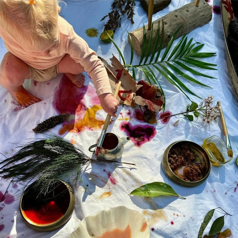 Child sitting on a white cloth surrounded by natural items, bright colored paint splatters, leaves, pine branches, a teapot, and other art supplies.