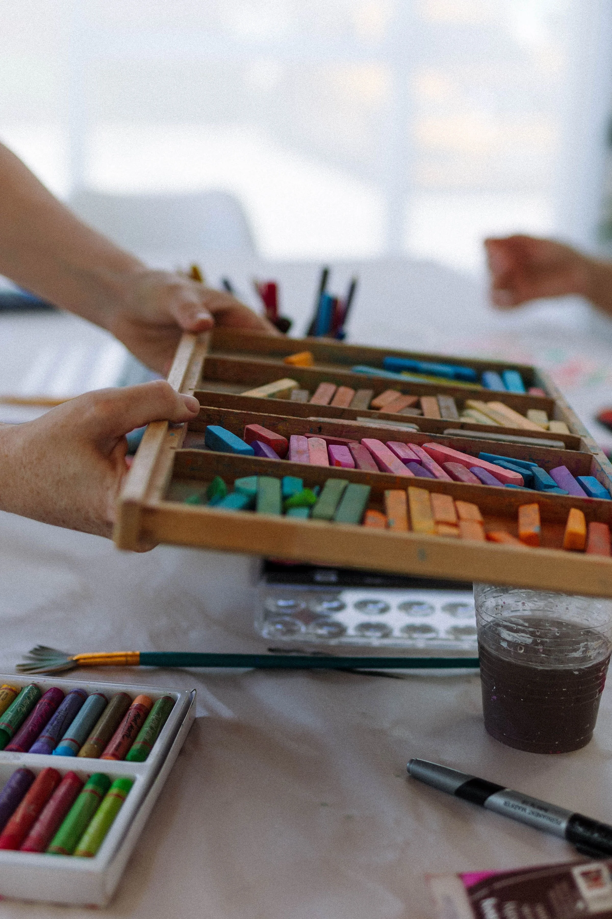 Close-up of a box of colorful chalks on a table, with hands reaching to pick a piece. In the background, there are more chalks and a pen on a white table. A cup of dark soda is visible to the side.