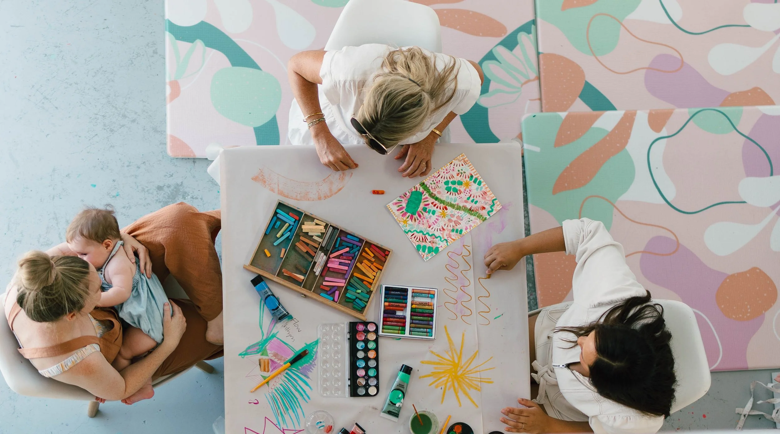 People at a table engaging in children's art activities with colorful chalk pastels, markers, and drawings, including abstract and nature-inspired designs.
