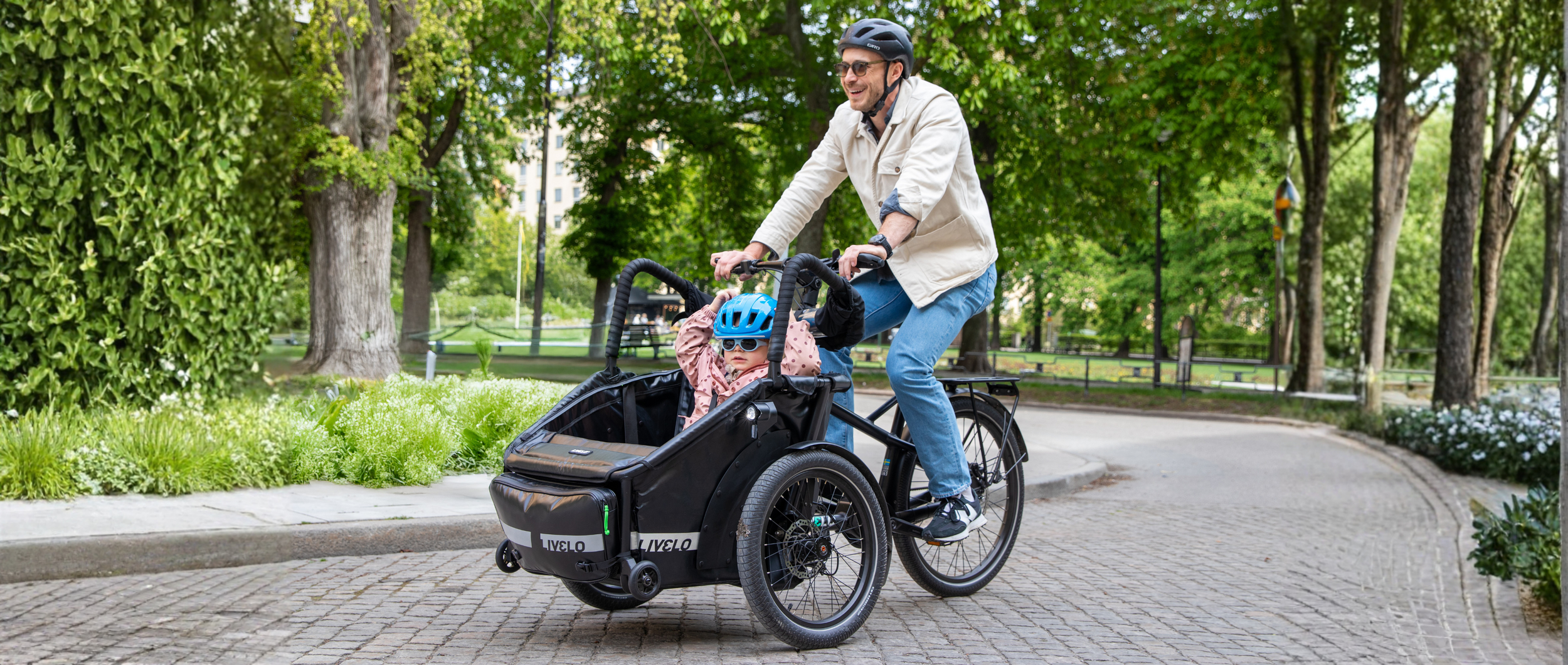 Pappa cyklar på en Livelo elassisterad lådcykel med sin dotter tryggt sittande i korgen i Stockholm, tessinparken.