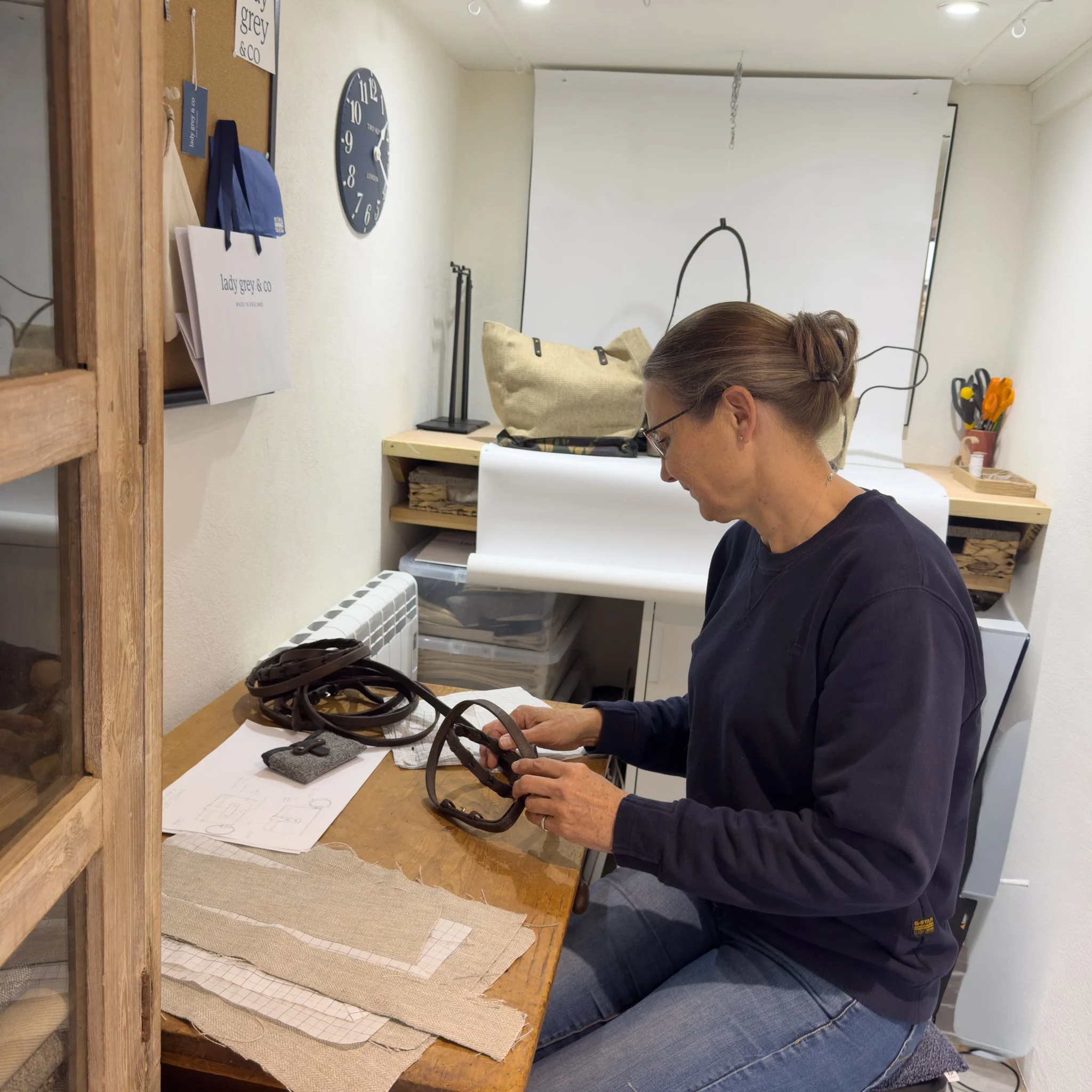 A woman with glasses and brown hair in a bun sitting at a wooden desk in a workshop or studio, examining a pair of glasses. The desk has fabric swatches, papers, and a small gray case. Behind her, there's a wall with a corkboard, a blue clock, and some shelves with supplies.