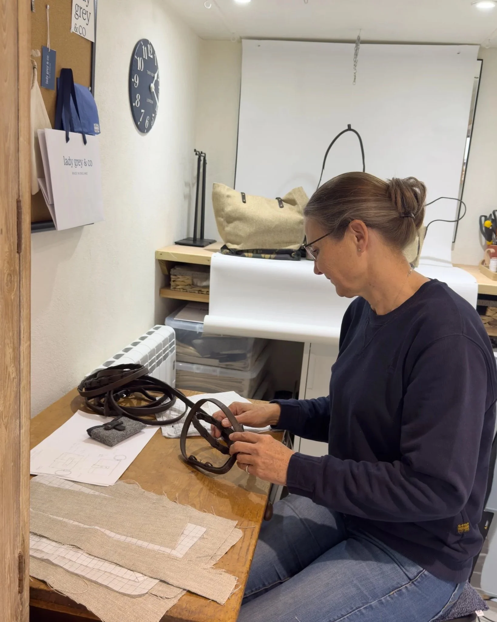 A woman with glasses and a bun working at a sewing or craft table with fabric swatches, patterns, and leather straps, in a craft room.