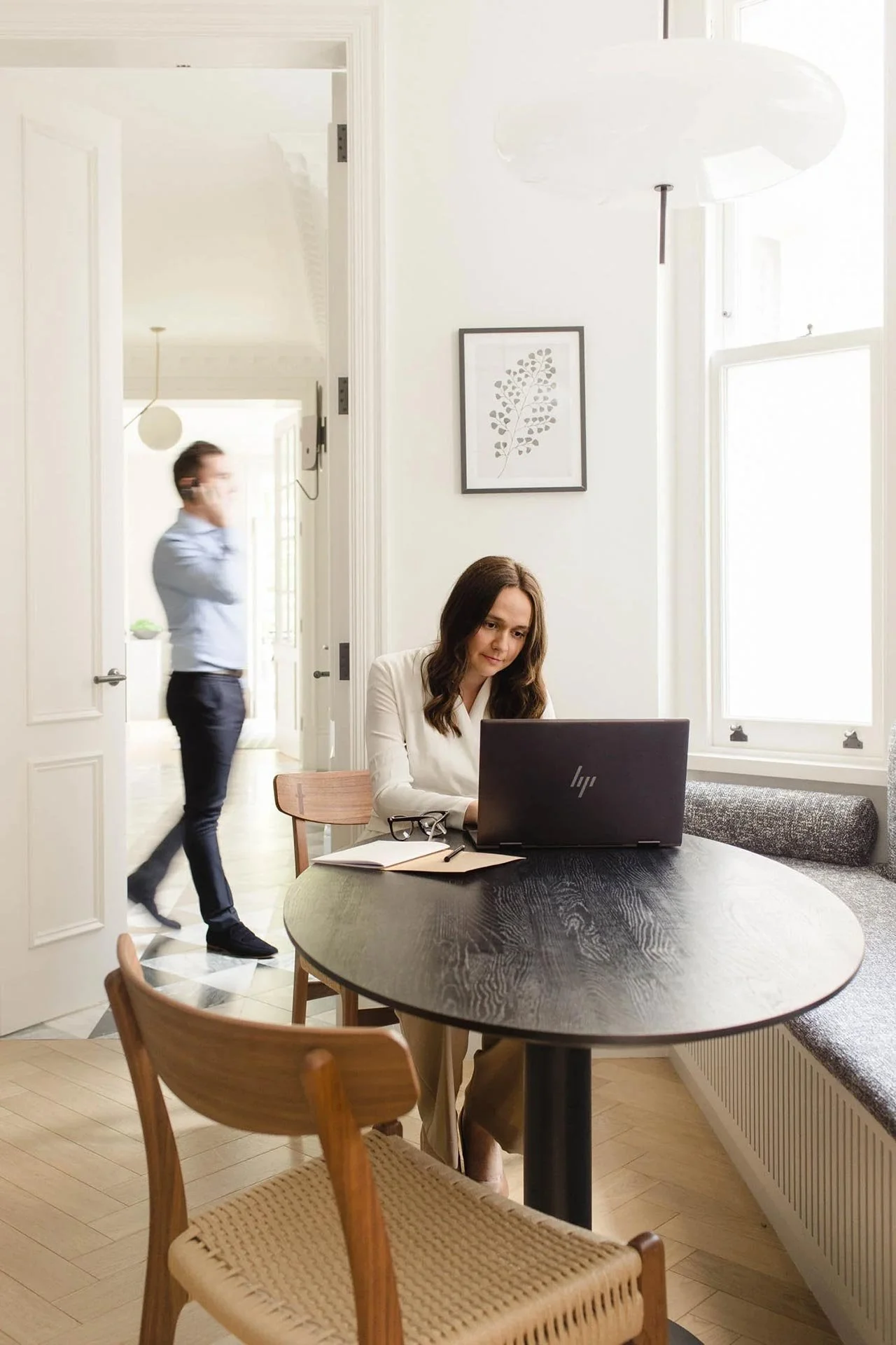 Woman in white top and fawn trousers sitting at a laptop with a man on a phone in background at a London accountancy practice