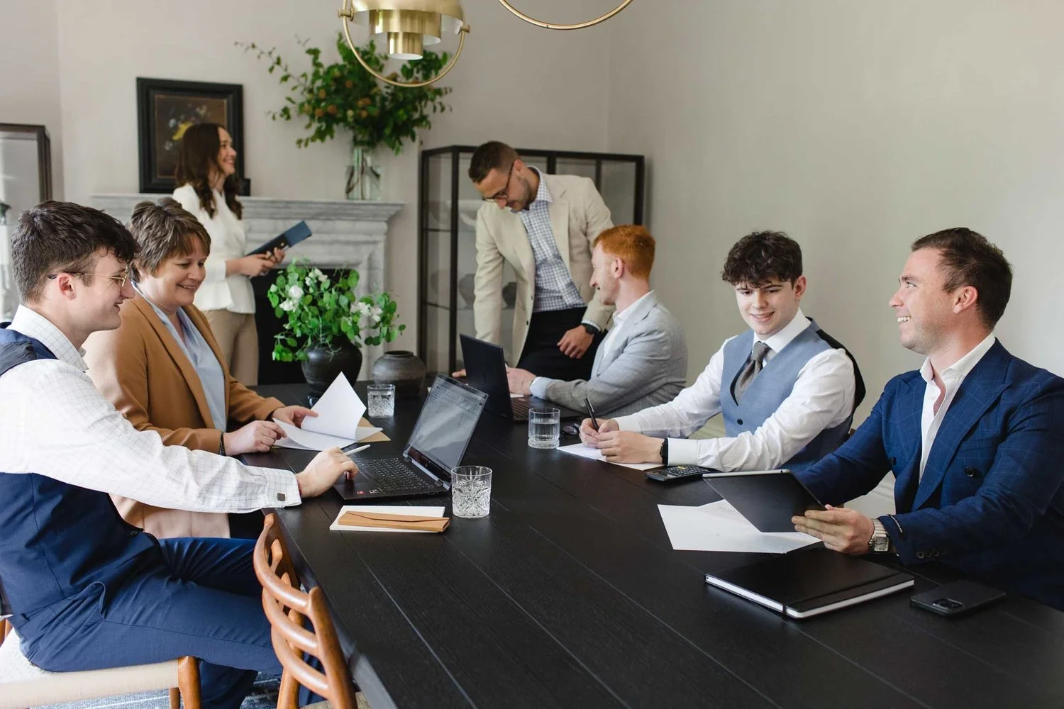 Seven accountants in smart attire work together around a dark wooden boardroom table using laptops tablets and notebooks