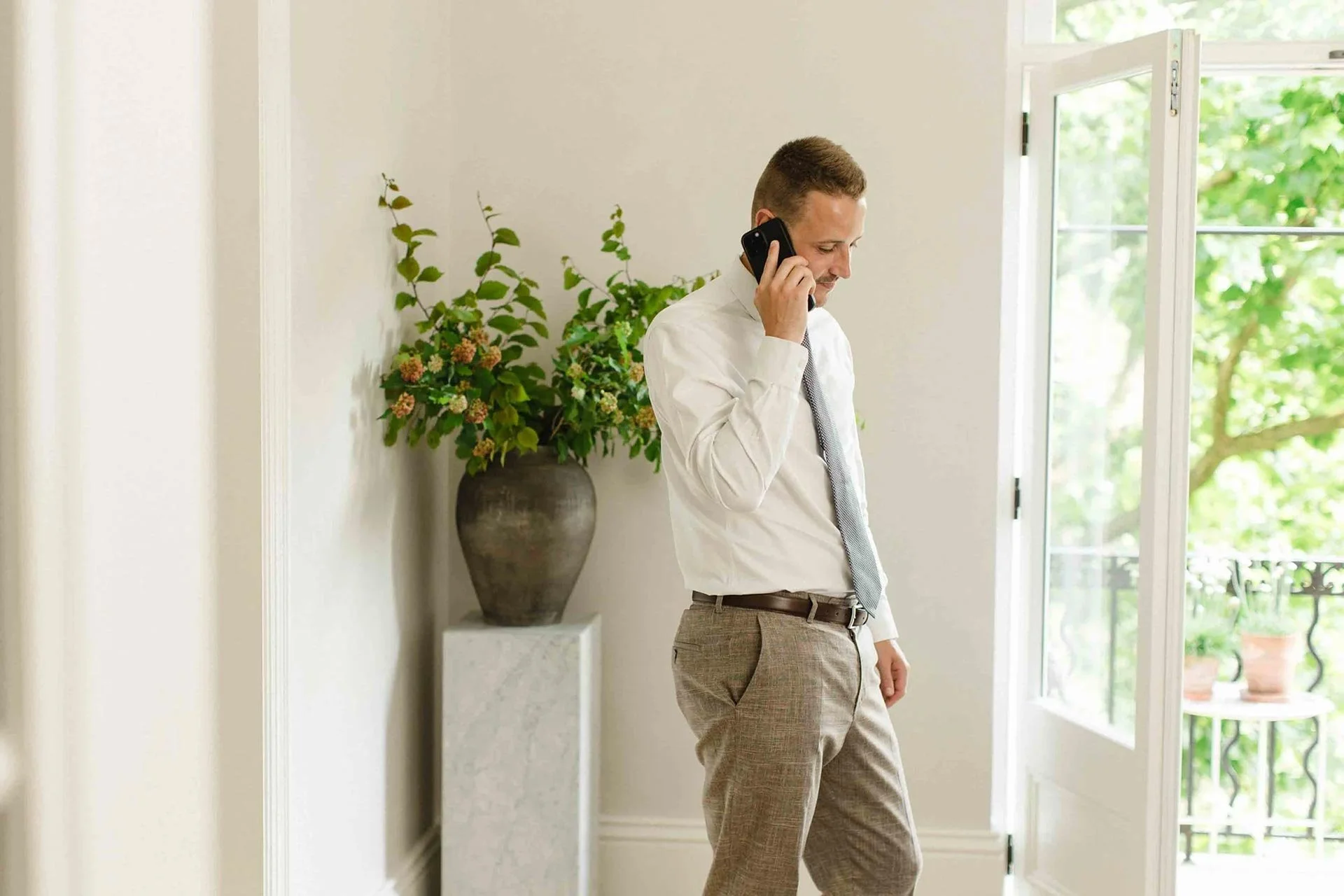 Man in white shirt and tie on mobile phone in light-filled room with plant on a marble pedestal and veranda in background