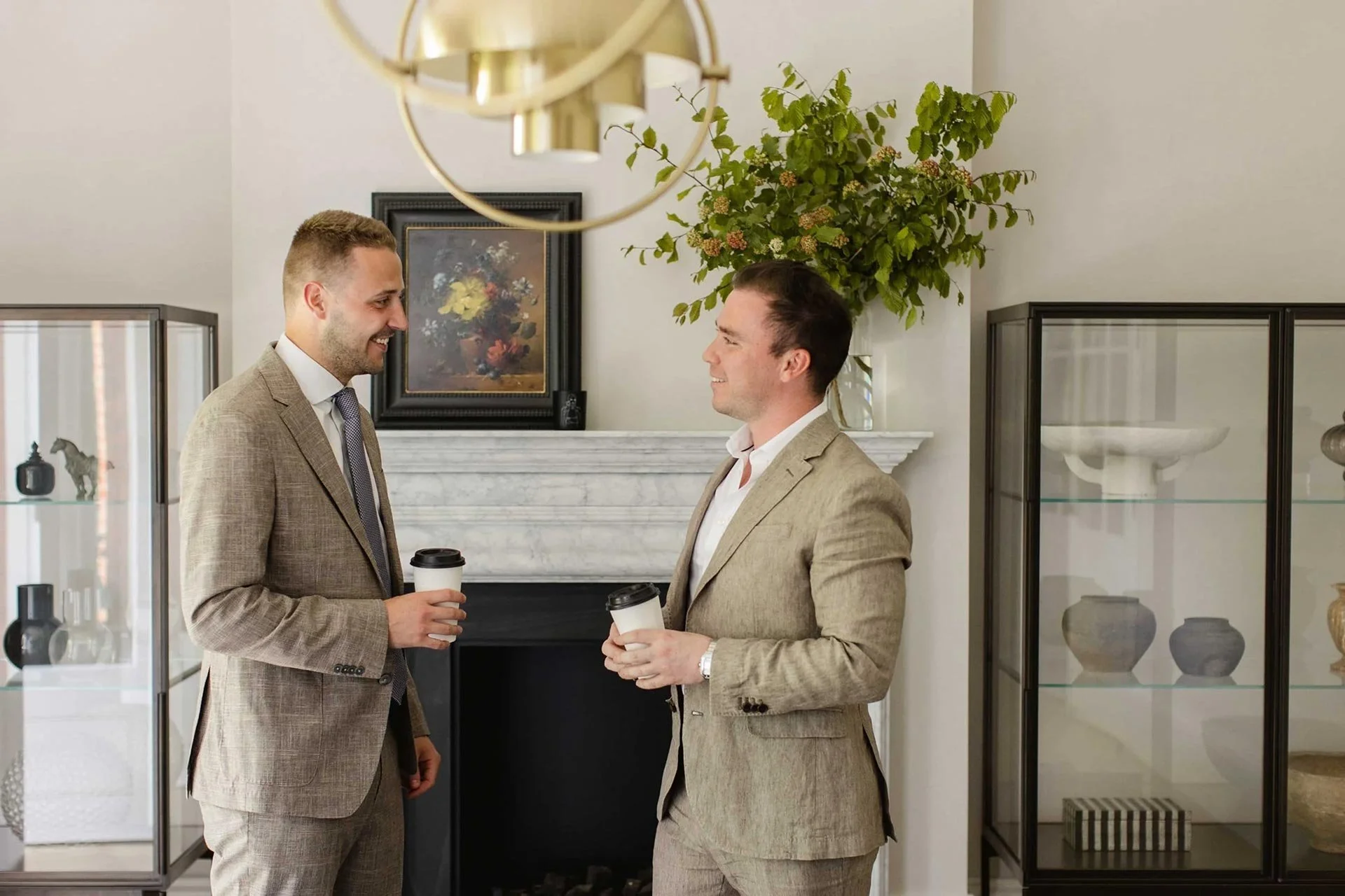 Two men in pale summer suits with open white shirts talking over a coffee in front of a fireplace at a London accountancy