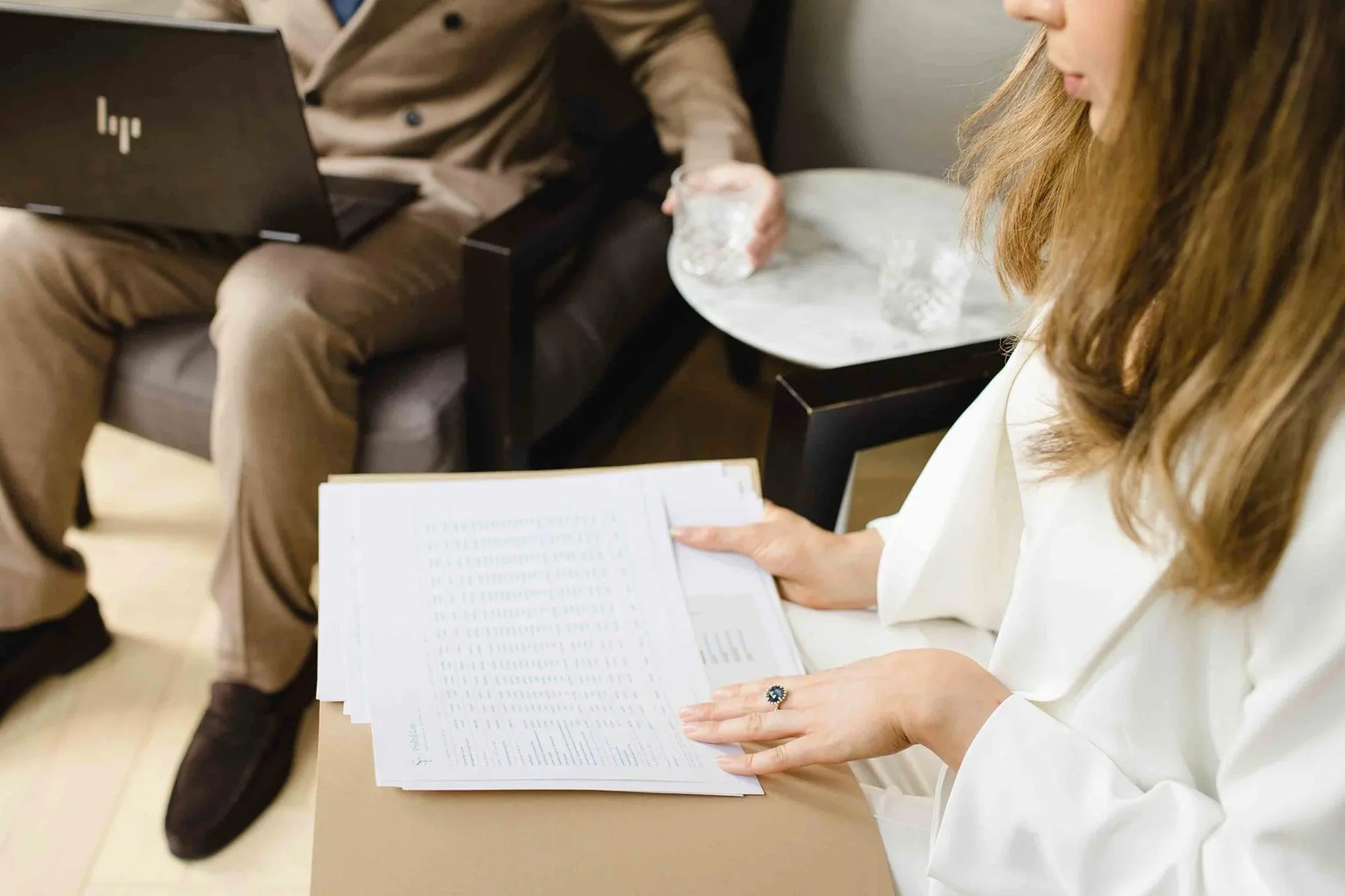 Woman with brown hair, white blazer suit with a blue ring reading financial information with man in brown suit and a laptop