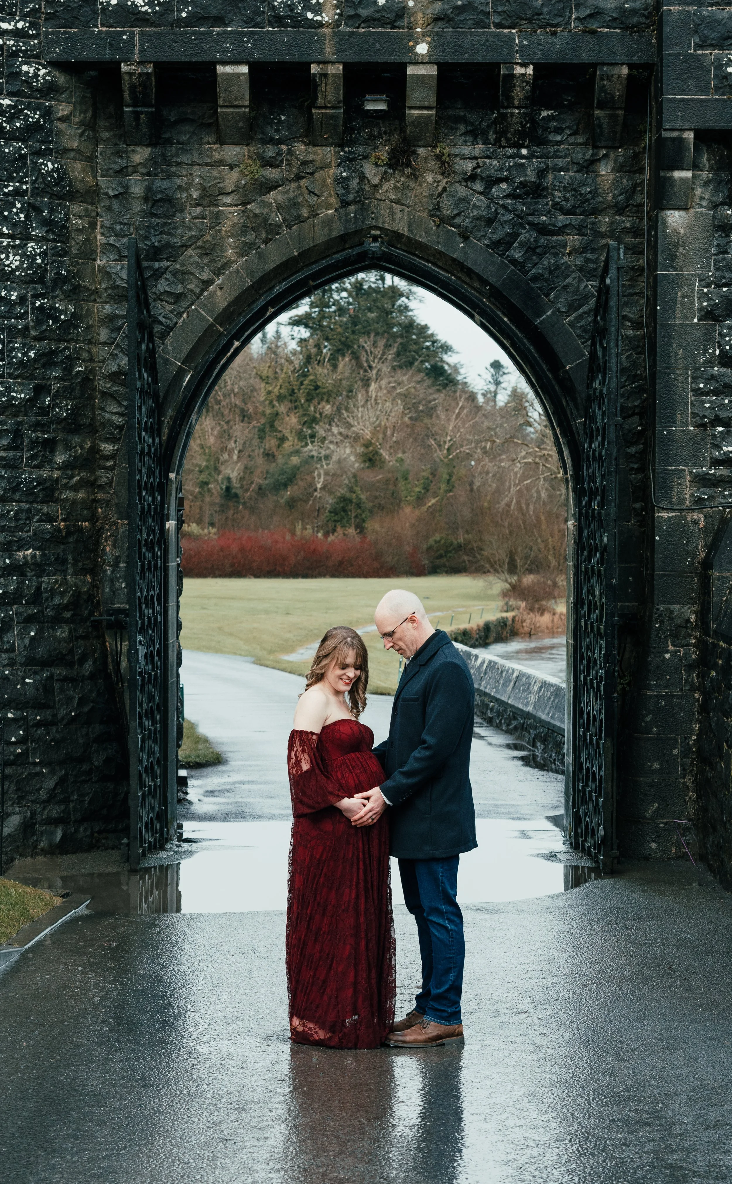 A couple, a pregnant woman in a red lace dress and a man in a dark coat and jeans, holding hands and looking at her baby bump under a stone archway on a rainy day.