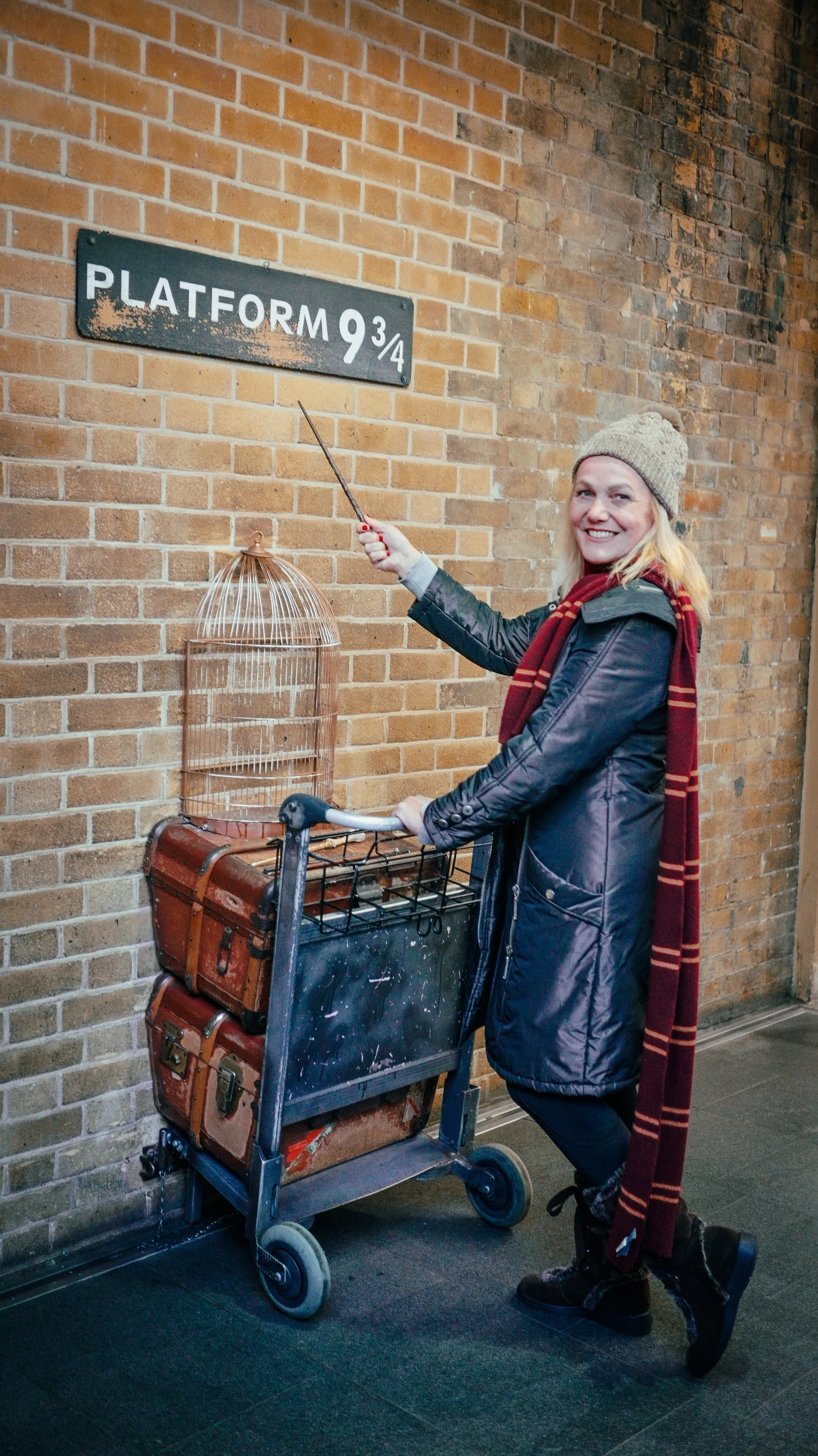 A woman dressed in a black coat, gray beanie, and red plaid scarf standing next to a luggage cart with vintage suitcases on it. She is smiling and pointing at a sign that says 'Platform 9 3/4' against a brick wall, referencing the Harry Potter series