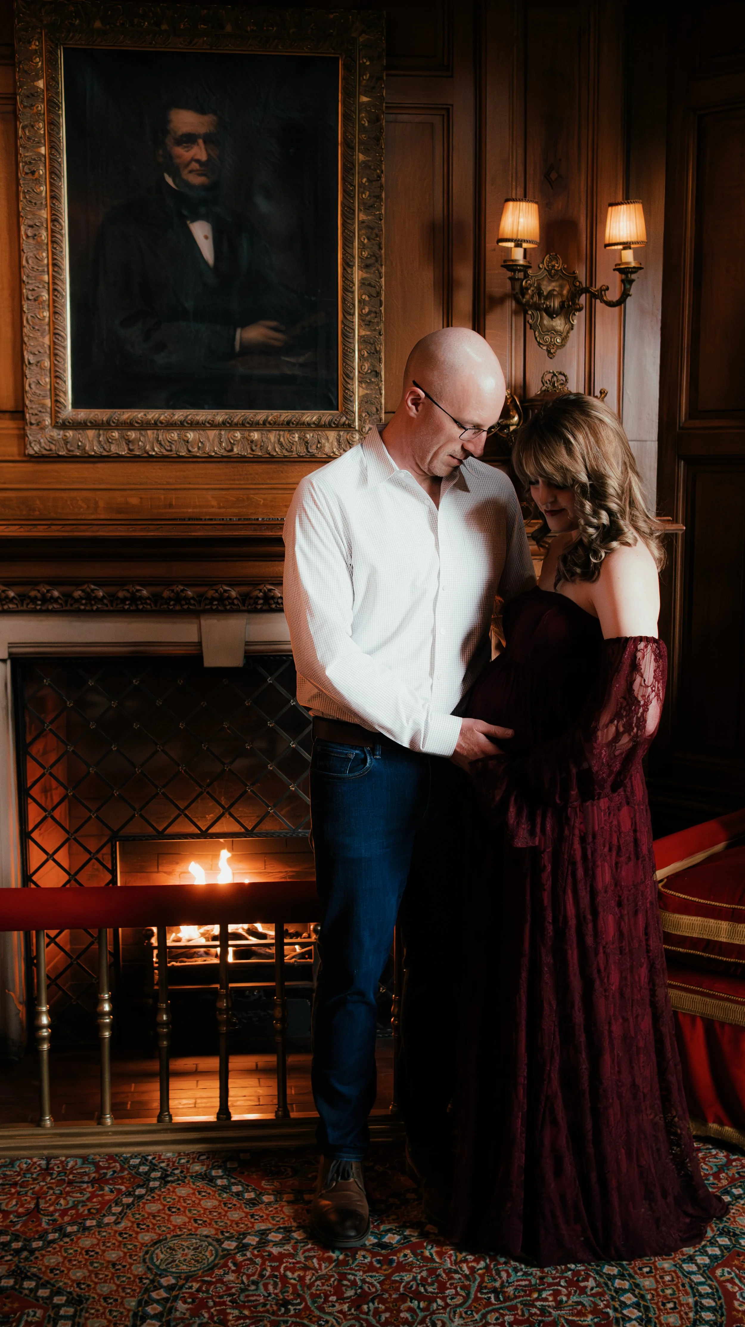 A man and a woman share a tender moment in front of a fireplace in a wood-paneled room. The man, wearing glasses and a white shirt, gently touches the woman's pregnant belly. The woman wears a dark red, lace dress and looks lovingly at her belly. A p