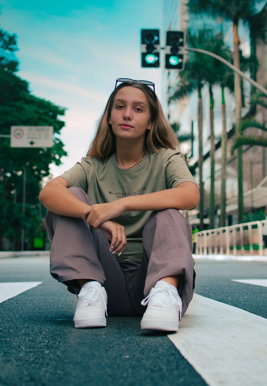 Young woman sitting on a pedestrian crosswalk on a city street during daytime, with traffic lights and tall buildings in the background.