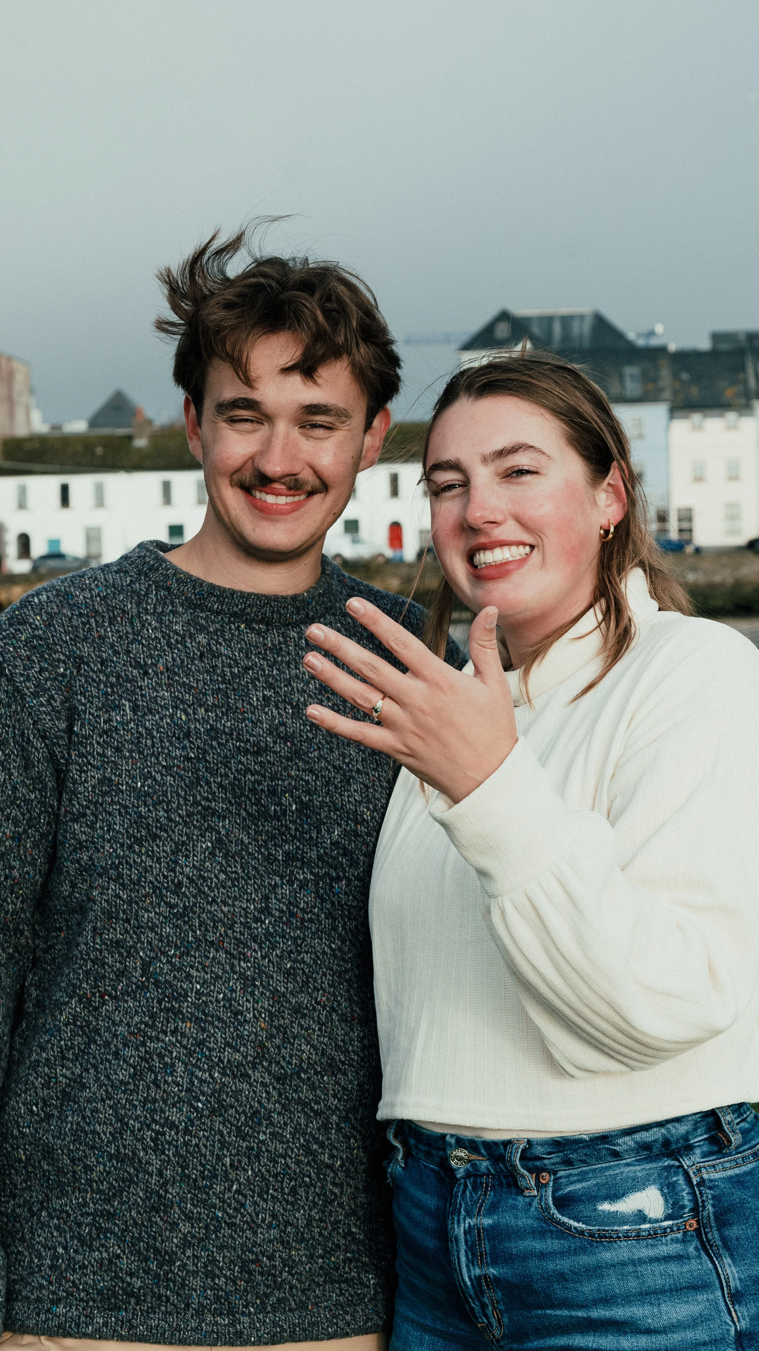 A smiling young couple standing outdoors with a row of white buildings in the background, the woman showing off an engagement ring on her left hand.