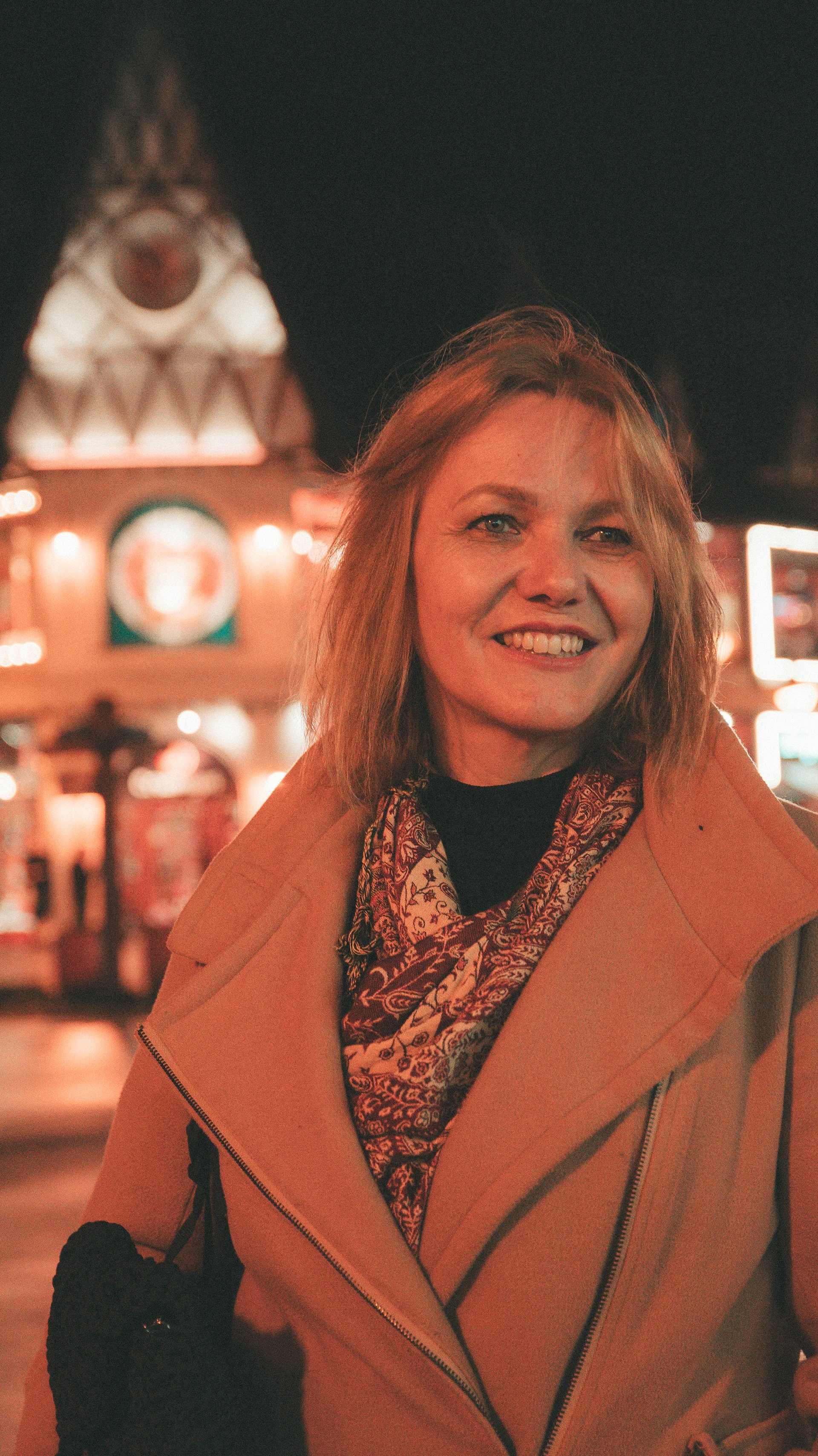 A woman with shoulder-length red hair smiling at night in an urban area with illuminated signs and buildings in the background.