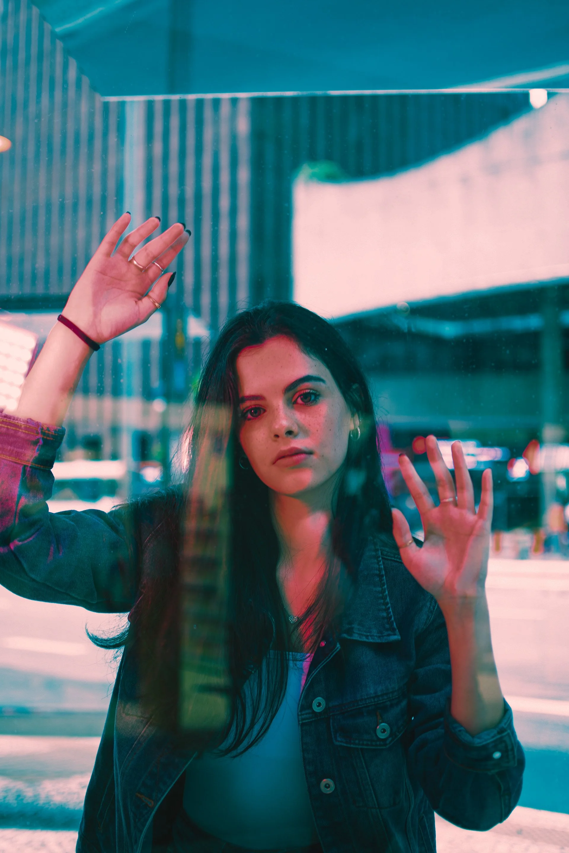 Young woman with black hair and pale skin looks through a glass window with colorful reflections, raising her hands to touch the glass in an urban setting.