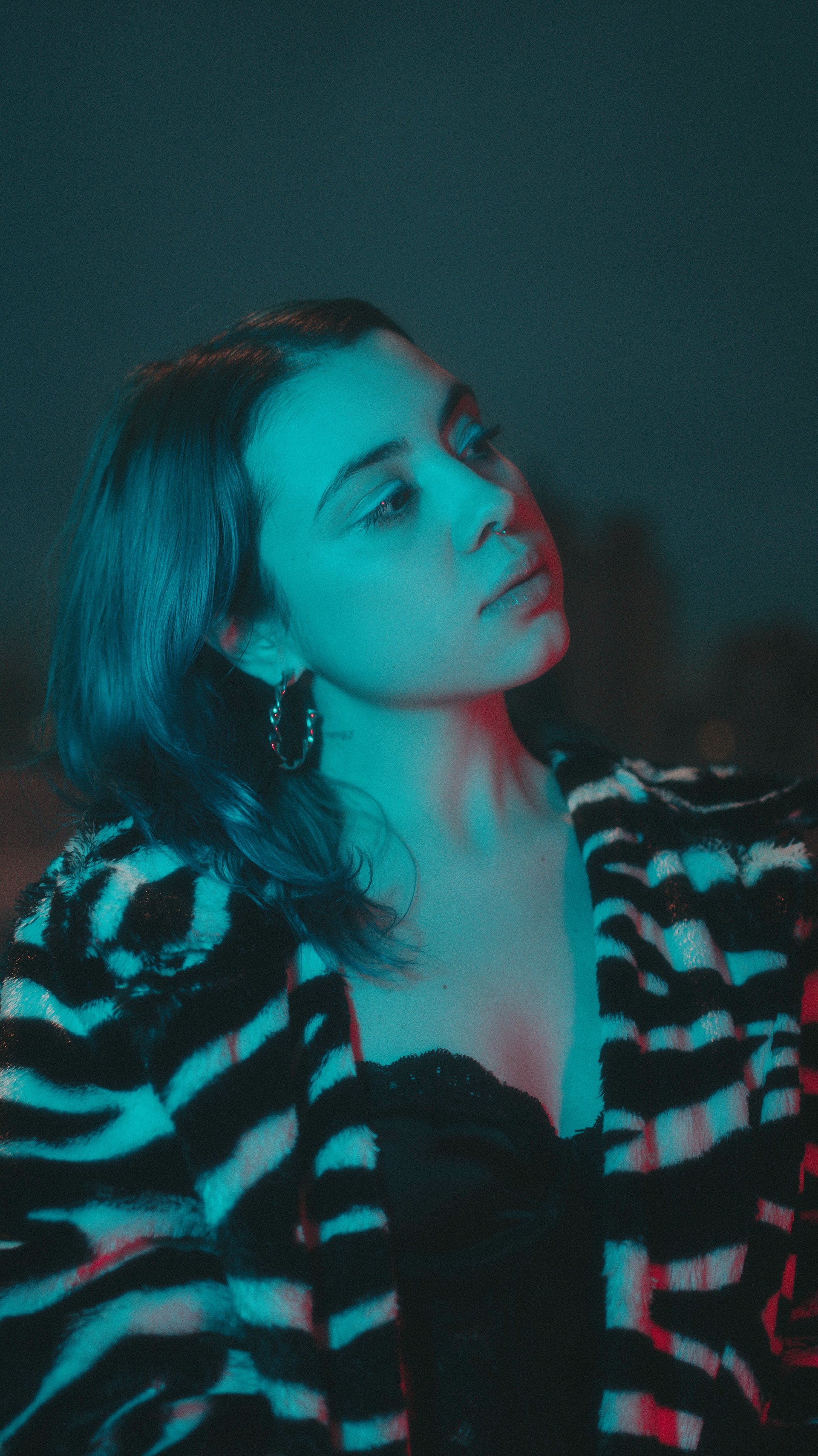 A young woman with shoulder-length dark hair and hoop earrings, wearing a black top with a laced neckline and a black and white striped jacket, is looking off to the side with a contemplative expression. She is illuminated by blue and red lighting ag