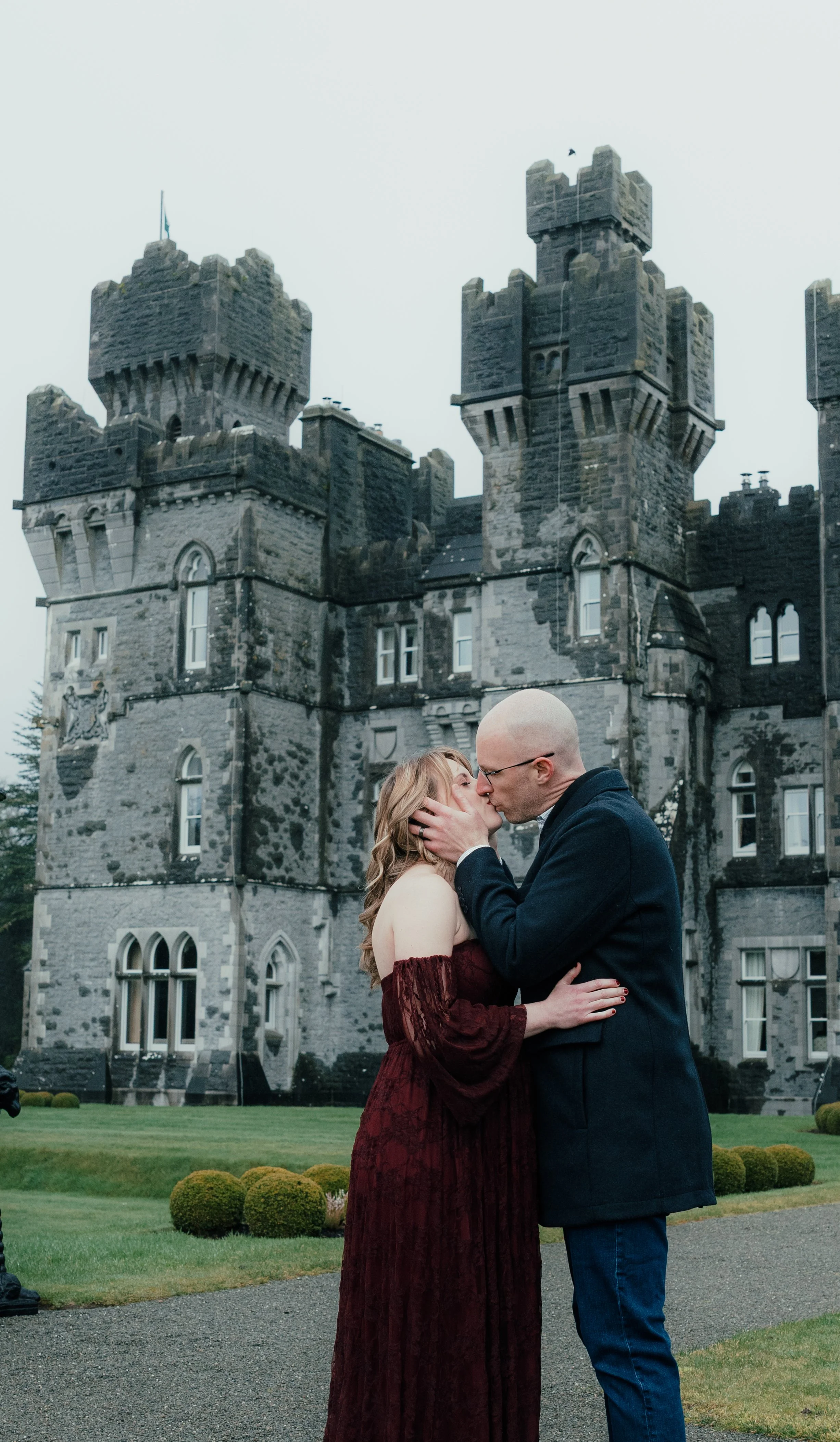 A couple kissing in front of a castle, with the woman wearing a strapless burgundy gown and the man in a dark coat and glasses.