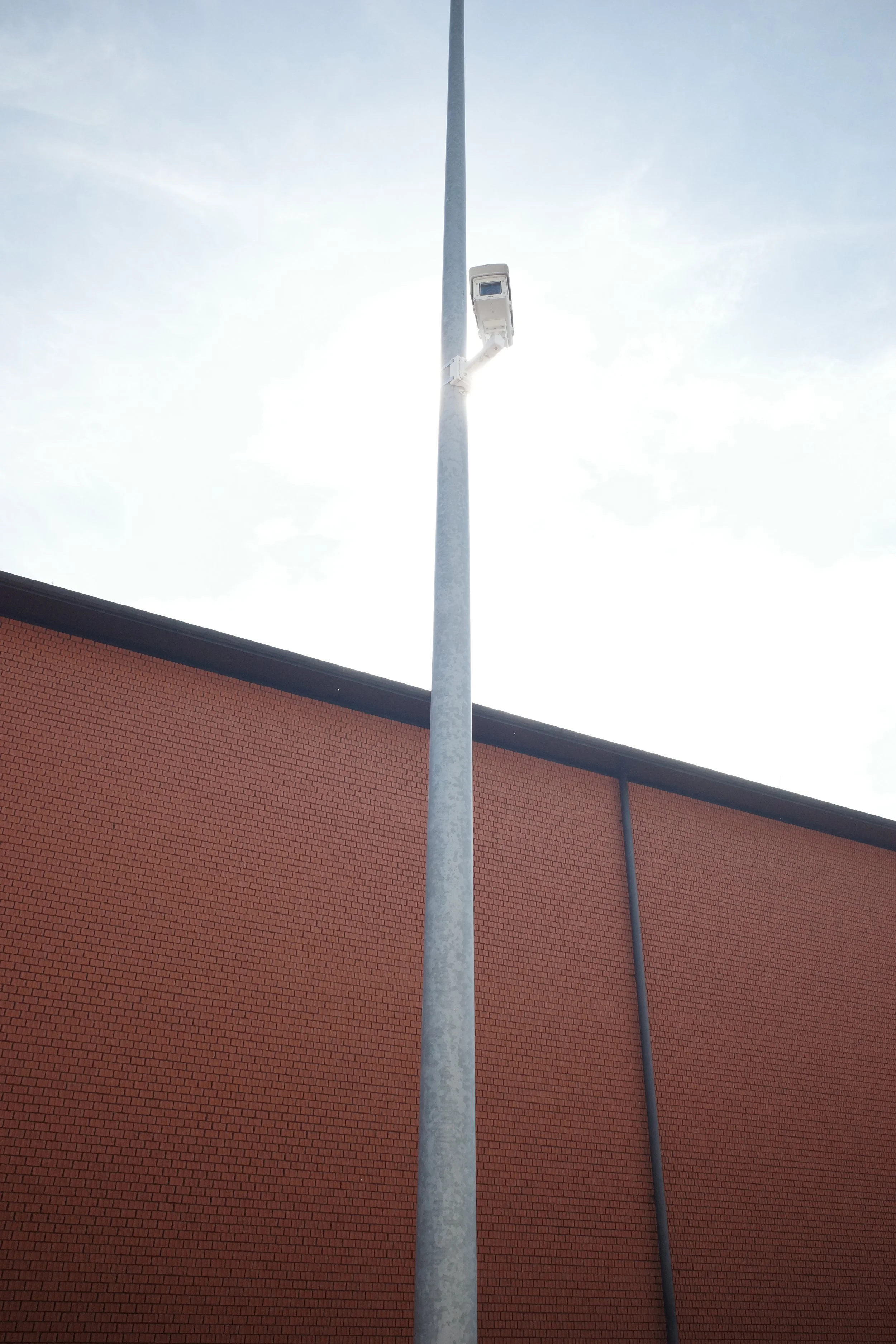 A tall streetlight pole with a surveillance camera attached, set against a brick building and cloudy sky.