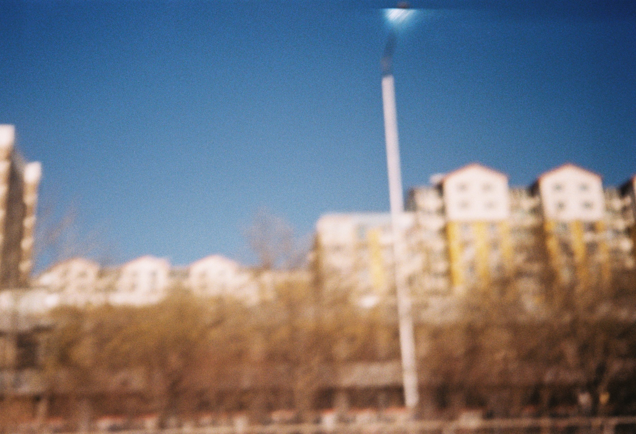Out-of-focus image of a cityscape featuring a blue sky, apartment buildings, and a streetlamp.