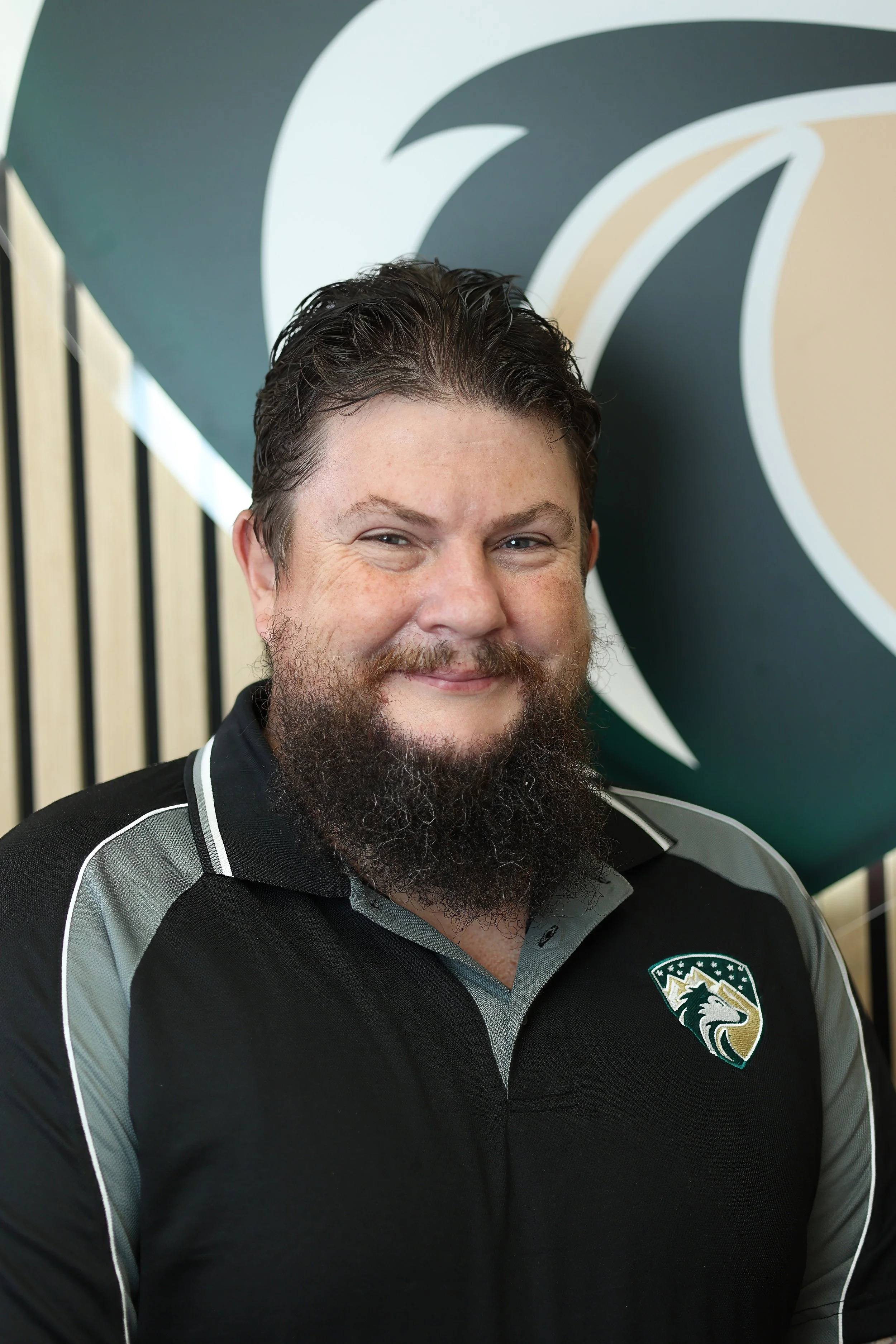 A man with dark hair, a beard, and mustache smiling, wearing a black and gray sports shirt with a logo on the chest, standing in front of a large abstract wall art.