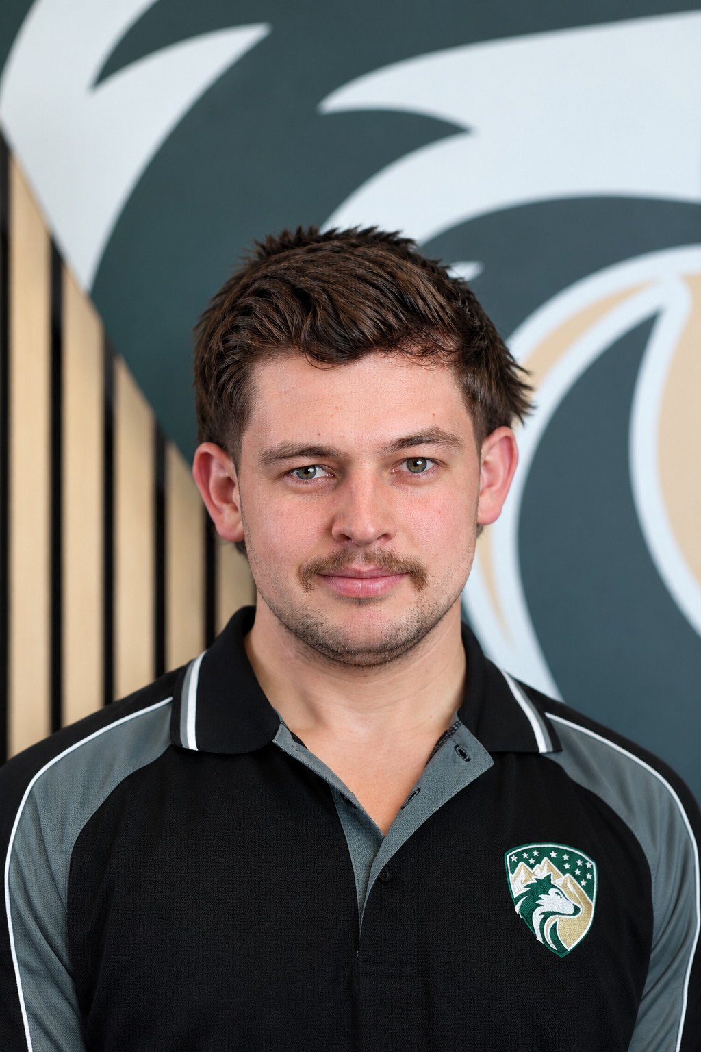 A young man with brown hair and a mustache dressed in a black and gray sports jersey with a wolf logo, standing in front of a stylized geometric wall mural.