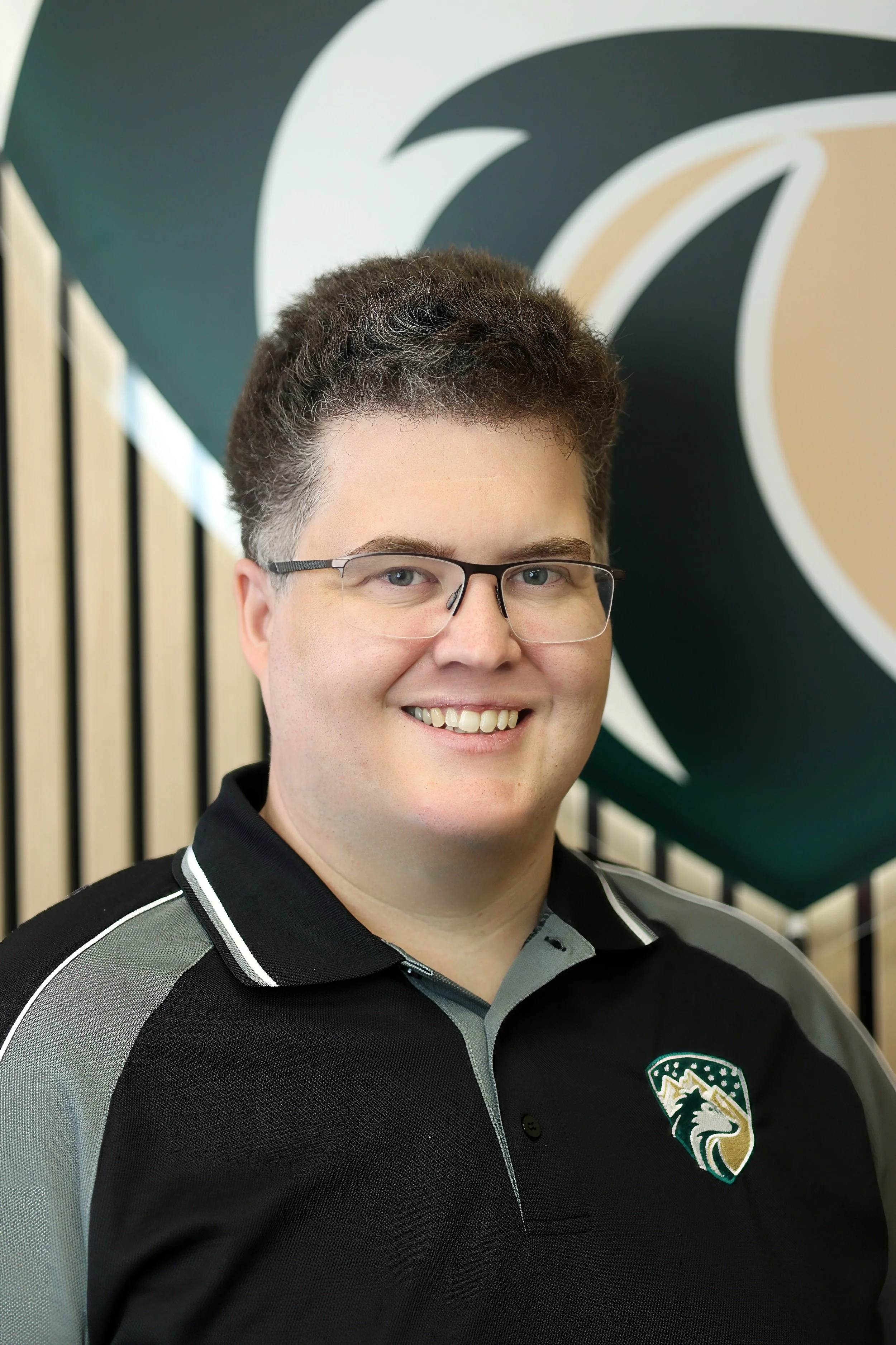 Man wearing glasses smiling, wearing a black and gray sports shirt with a logo on the chest, standing in front of abstract wall art with black, white, and tan patterns.