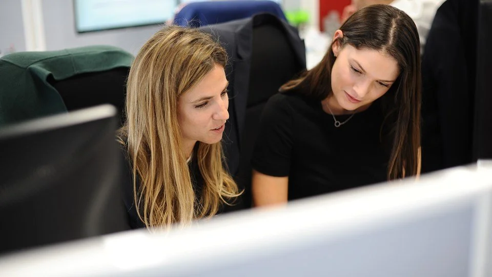 Two women seated at a desk, looking at a computer monitor, engaged in work or discussion in an office environment.
