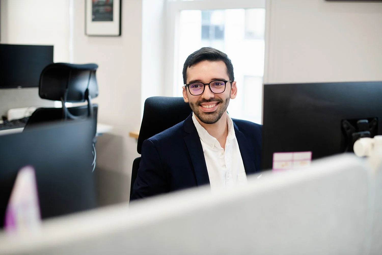 A smiling man with dark hair, beard, glasses, wearing a white shirt and dark blazer, sitting at a computer desk in an office.