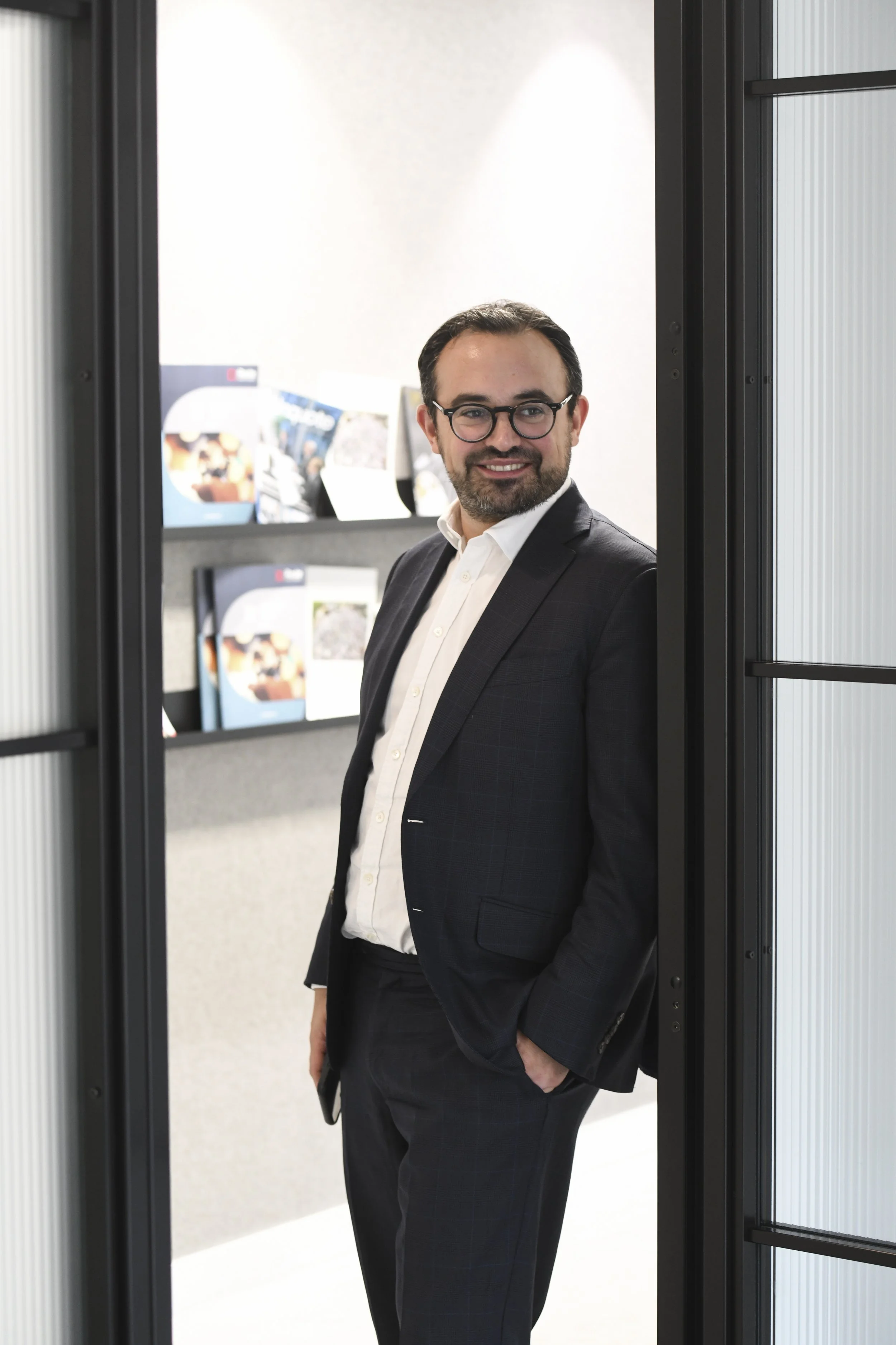 A man wearing glasses, a white shirt, and a dark suit, standing and smiling in an office hallway next to a glass wall, with shelves holding magazines or brochures in the background.