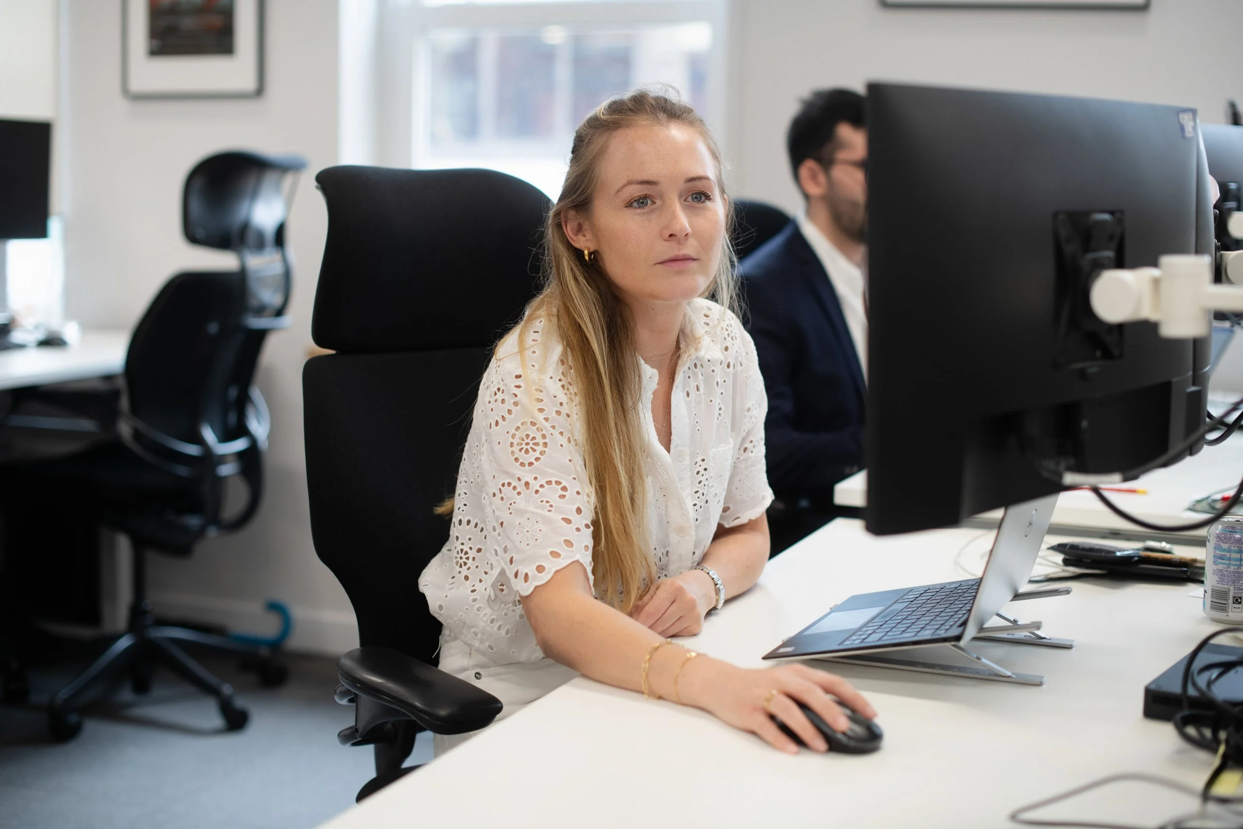 A woman with long blonde hair working at a desk in an office, using a computer mouse and looking at a monitor. She is wearing a white eyelet blouse and jewelry. Another man is working at a computer in the background.