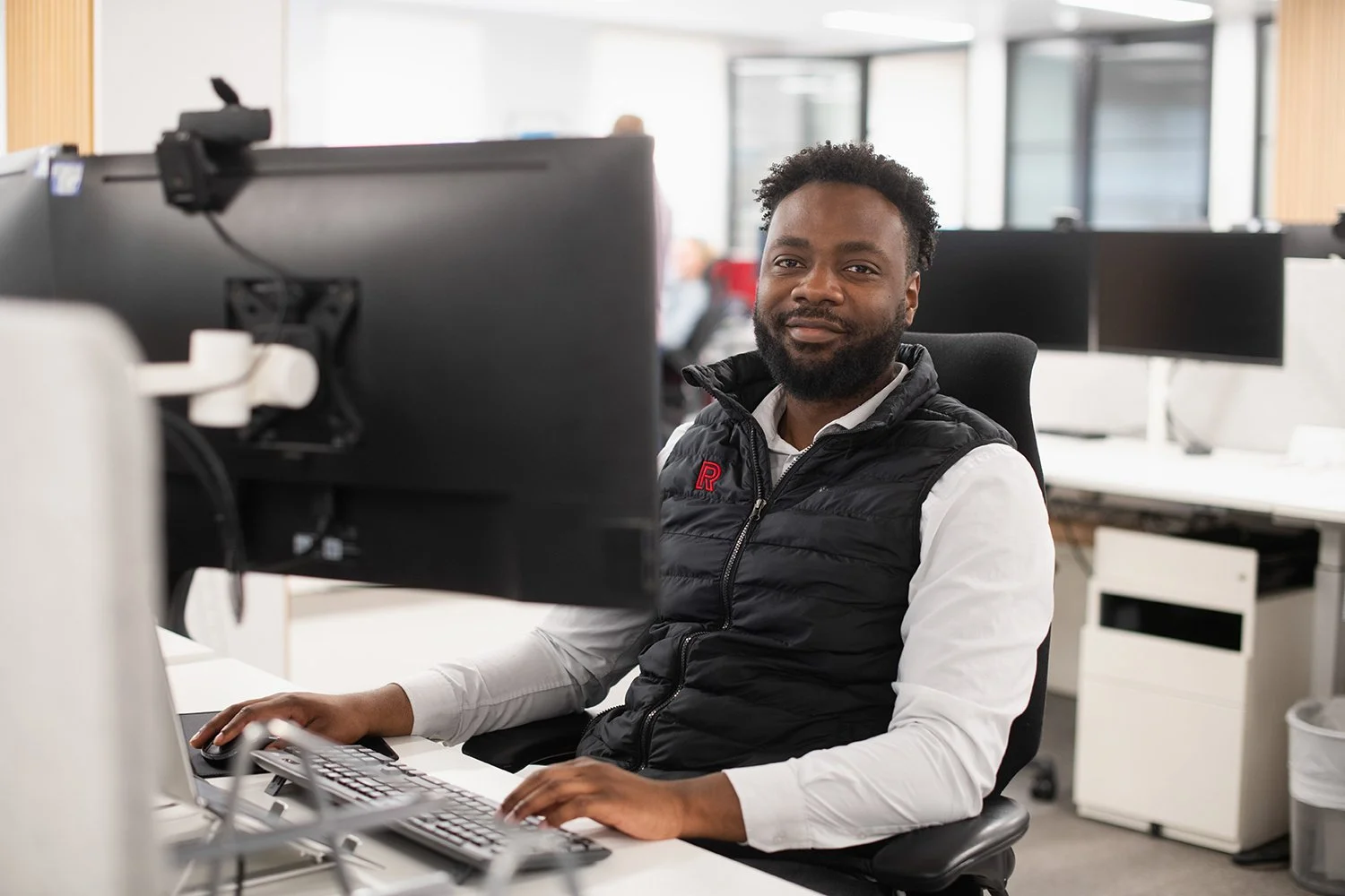 A man sitting at an office desk in front of a computer monitor, smiling at the camera.