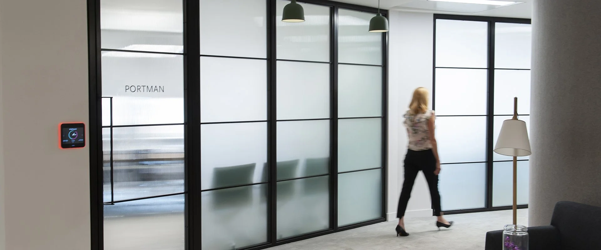 Woman walking past a glass door with frosted panels in a modern office building, labeled 'PORTMAN' on the door, with contemporary lighting and furniture.