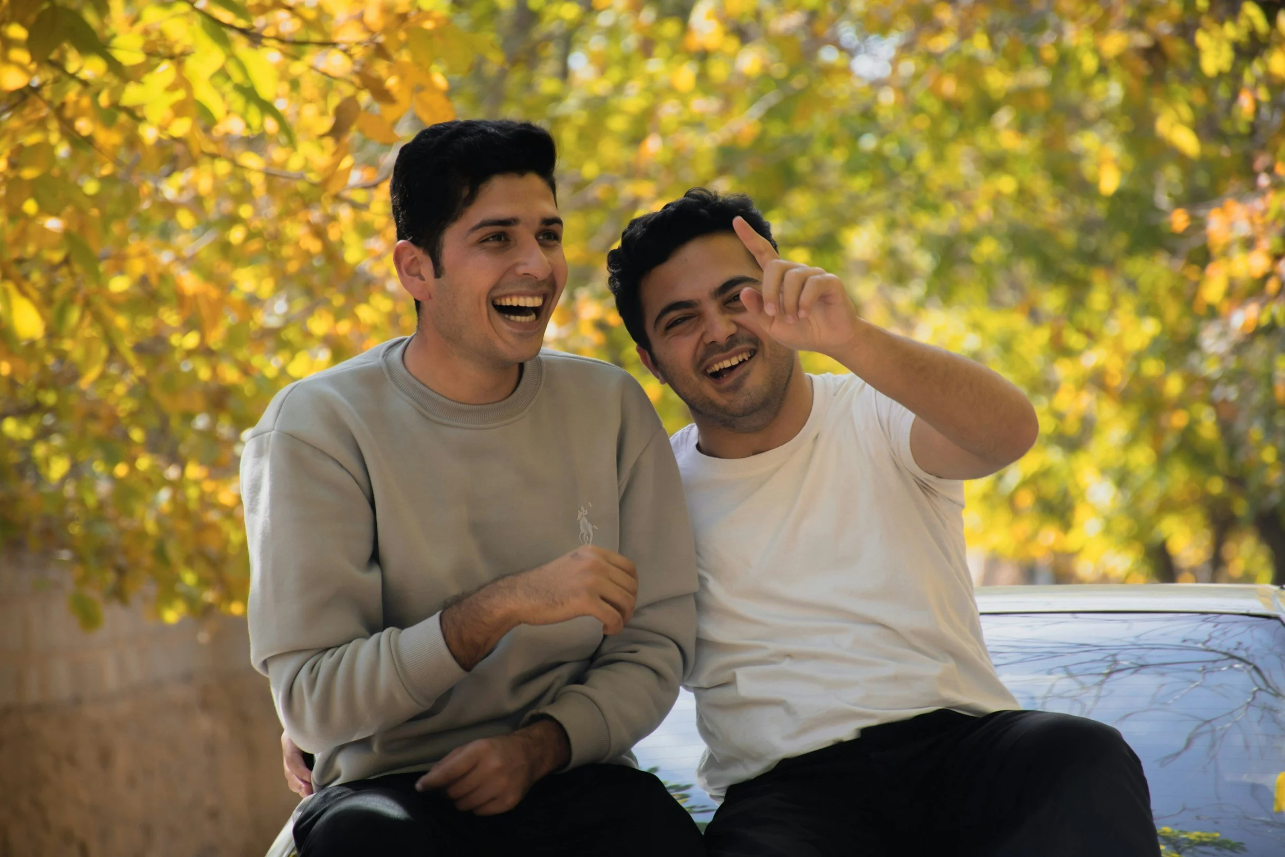 Two young men sitting outdoors near a body of water, laughing and enjoying each other's company during autumn with yellow and orange leaves in the background.