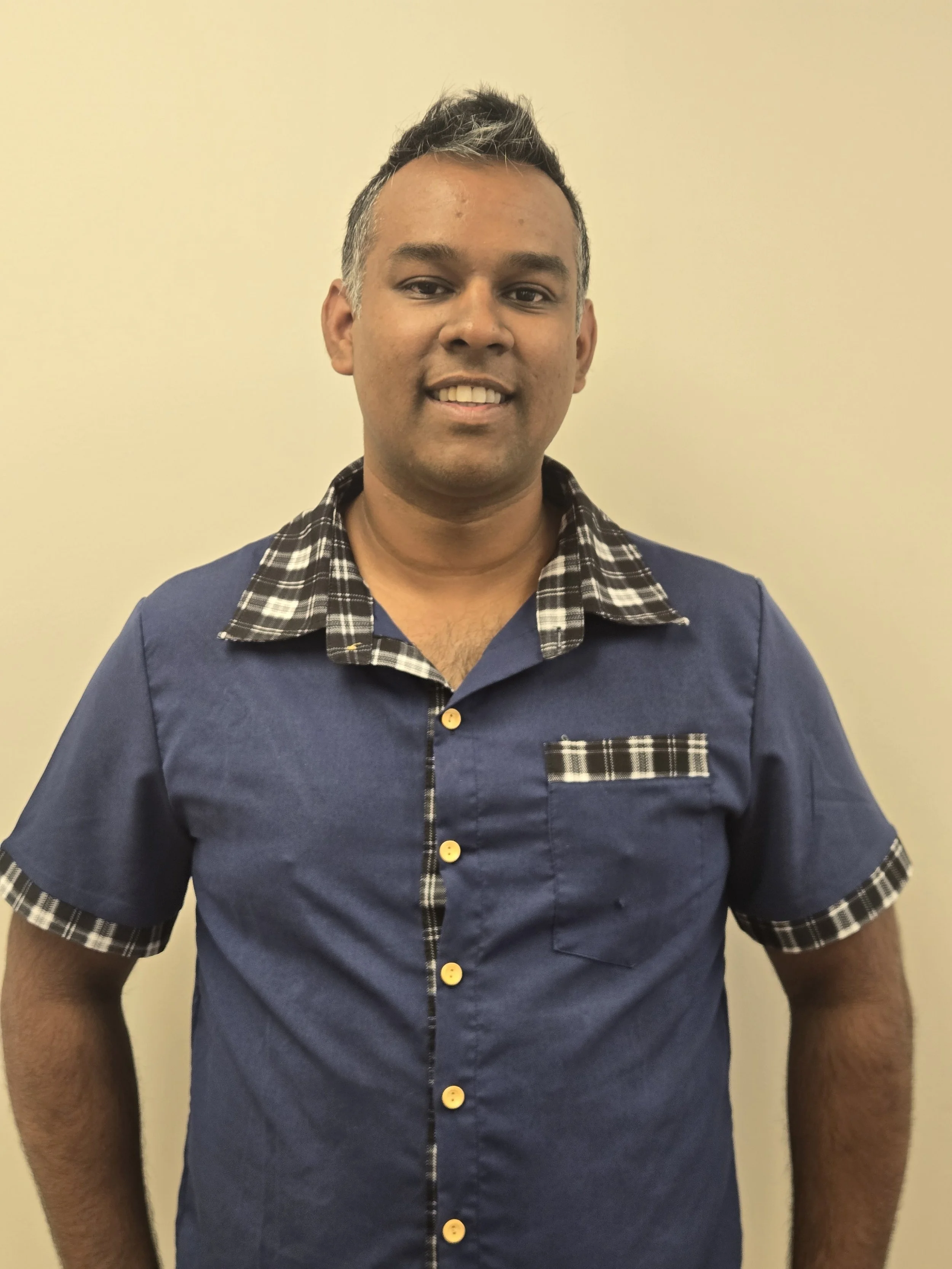 A man with short tapered hair with white highlights, wearing a navy blue shirt with a checkered collar and sleeve trim, standing against a plain beige wall.
