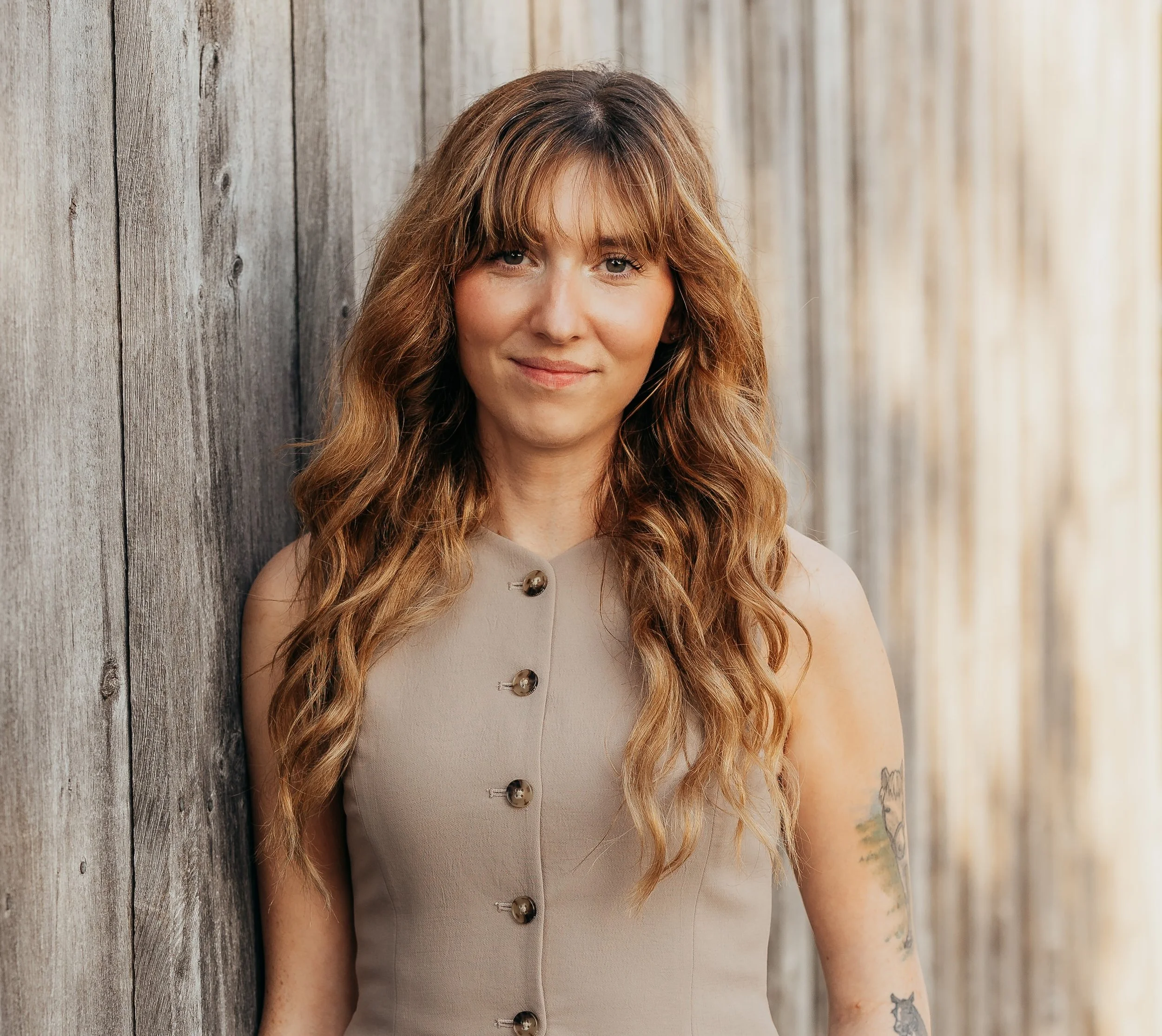 Front view of a woman with long wavy hair, wearing a beige sleeveless top with large buttons, standing against a weathered wooden wall.