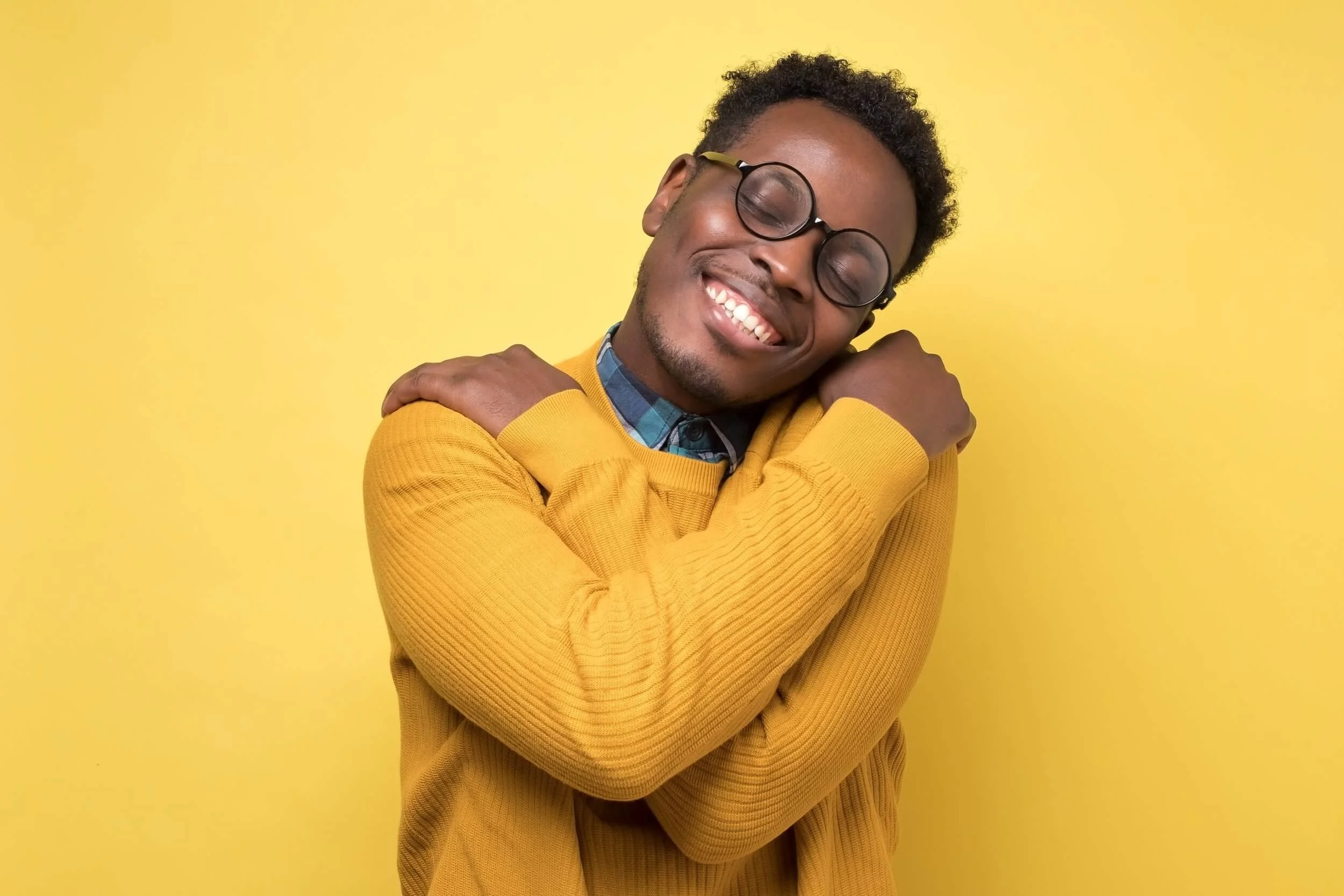 A young man with curly hair and glasses, smiling with closed eyes, hugging himself, wearing a yellow sweater against a yellow background.
