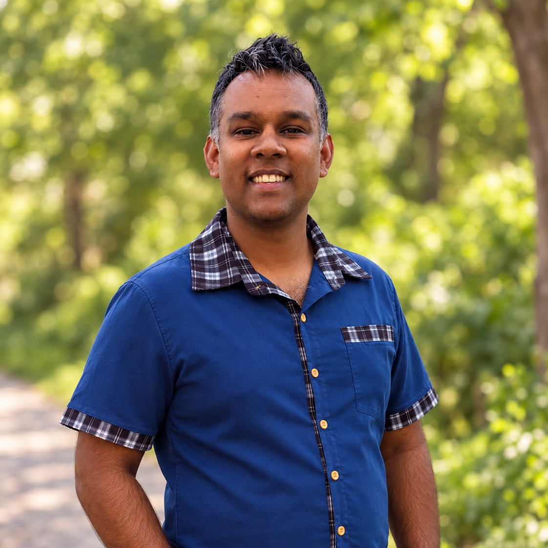 A smiling man with short black hair wearing a blue short-sleeved shirt with plaid accents, standing outdoors with green trees and sunlight in the background.
