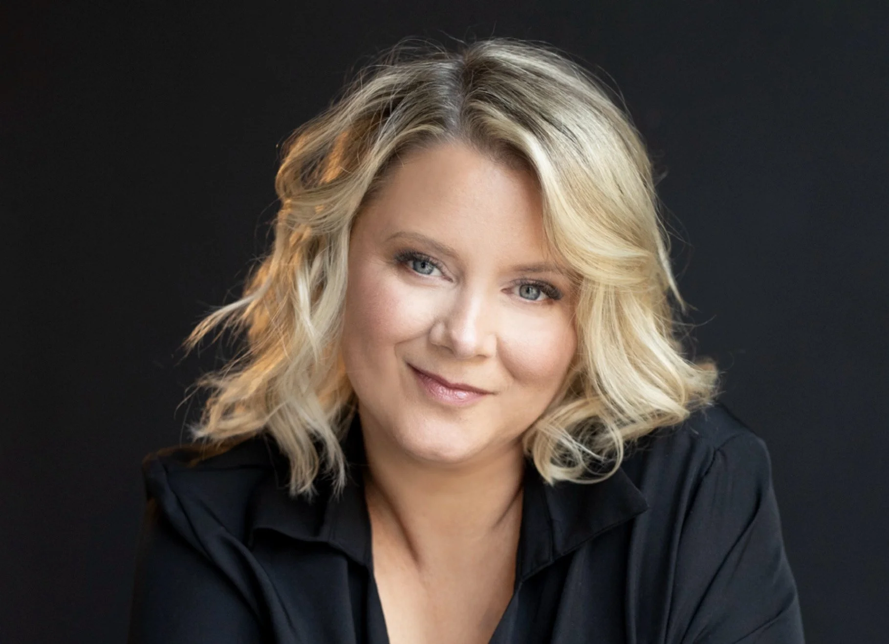 Close-up portrait of a blonde woman with wavy hair and blue eyes, smiling softly against a dark background.