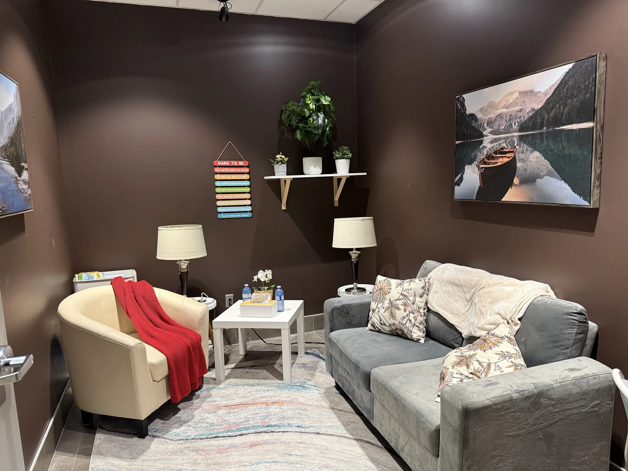A cozy living room with dark brown walls, a gray couch with floral pillows, a beige armchair with a red throw blanket, and a white coffee table with water bottles and a small plant. There are two lamps on side tables and framed landscape pictures on the walls, including a lake with mountains. A carpet with pastel colors covers part of the tiled floor.