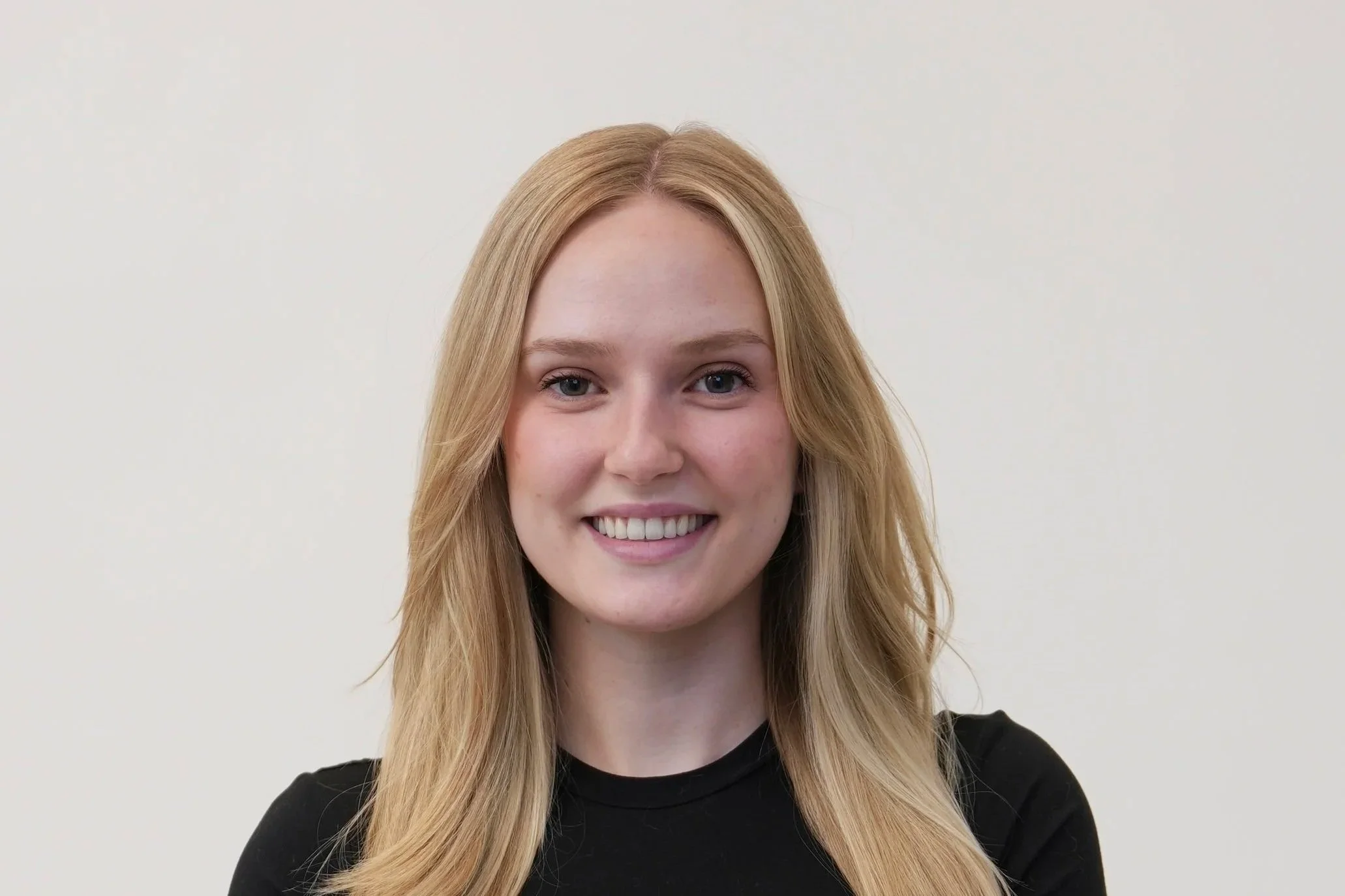 Close-up of a young woman with long blonde hair, smiling, wearing a black shirt against a plain white background.