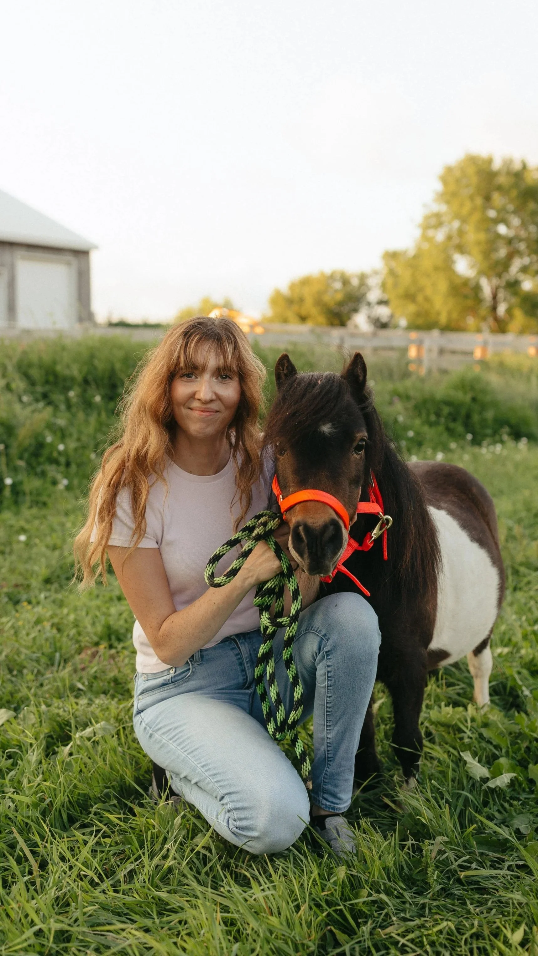 A woman with long, wavy red hair kneeling on green grass, holding a small black and white pony with an orange halter and green and black lead rope, outdoors in a rural setting during daytime.
