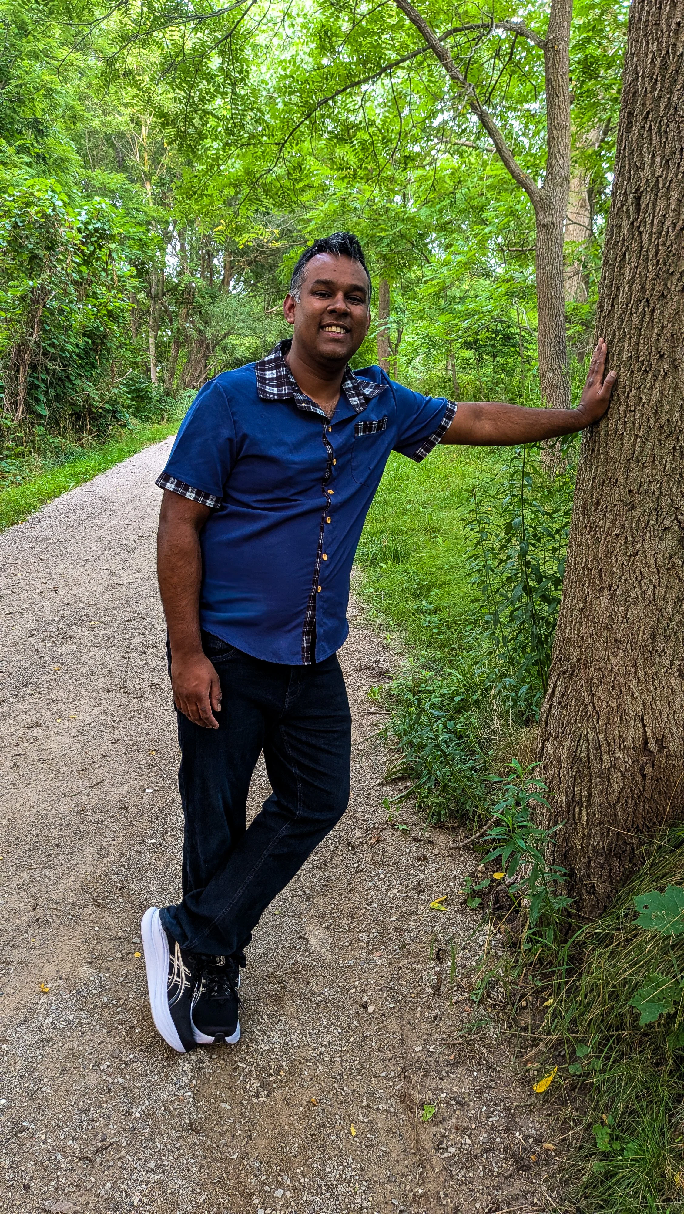 A man in a blue shirt with plaid accents, black pants, and black sneakers with white soles, standing on a dirt path in a green, wooded area. He is touching a tree with one hand and smiling at the camera.