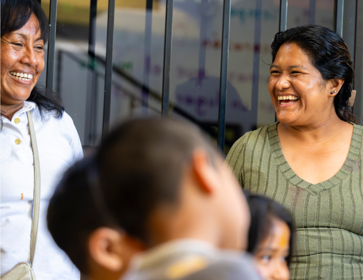 A group of women smiling and talking together in a community setting, with children in the foreground, conveying connection, collaboration, and shared care.