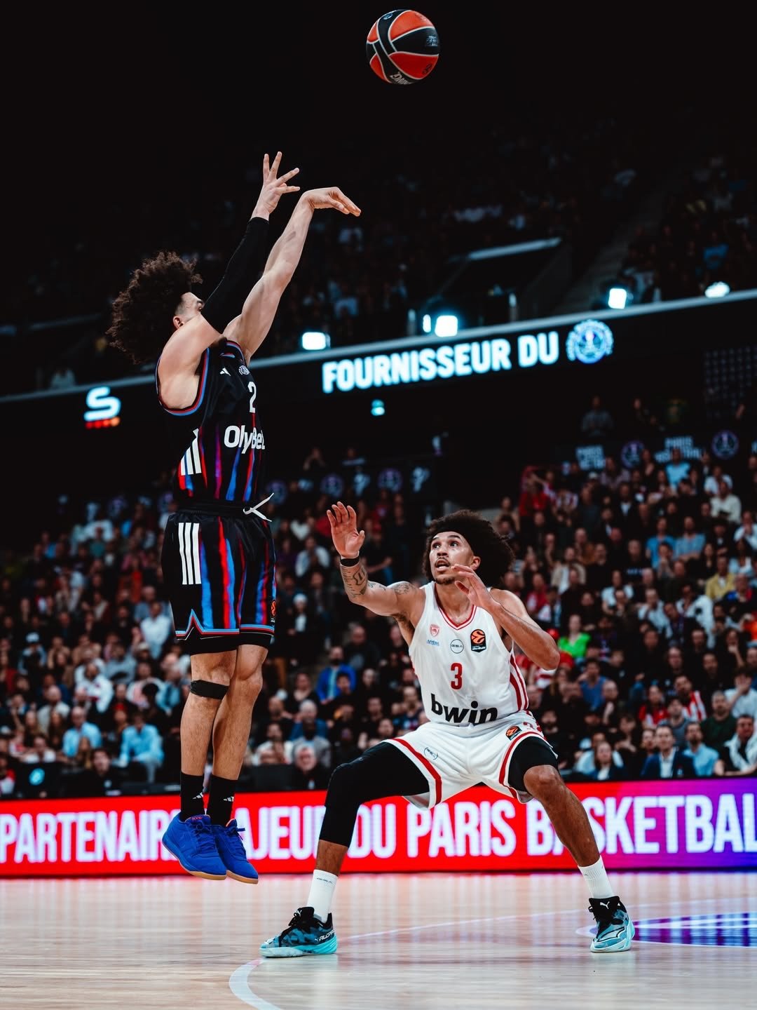 Two basketball players during a game, one jumping to shoot and the other crouched defensively, with a crowd of spectators in the background.