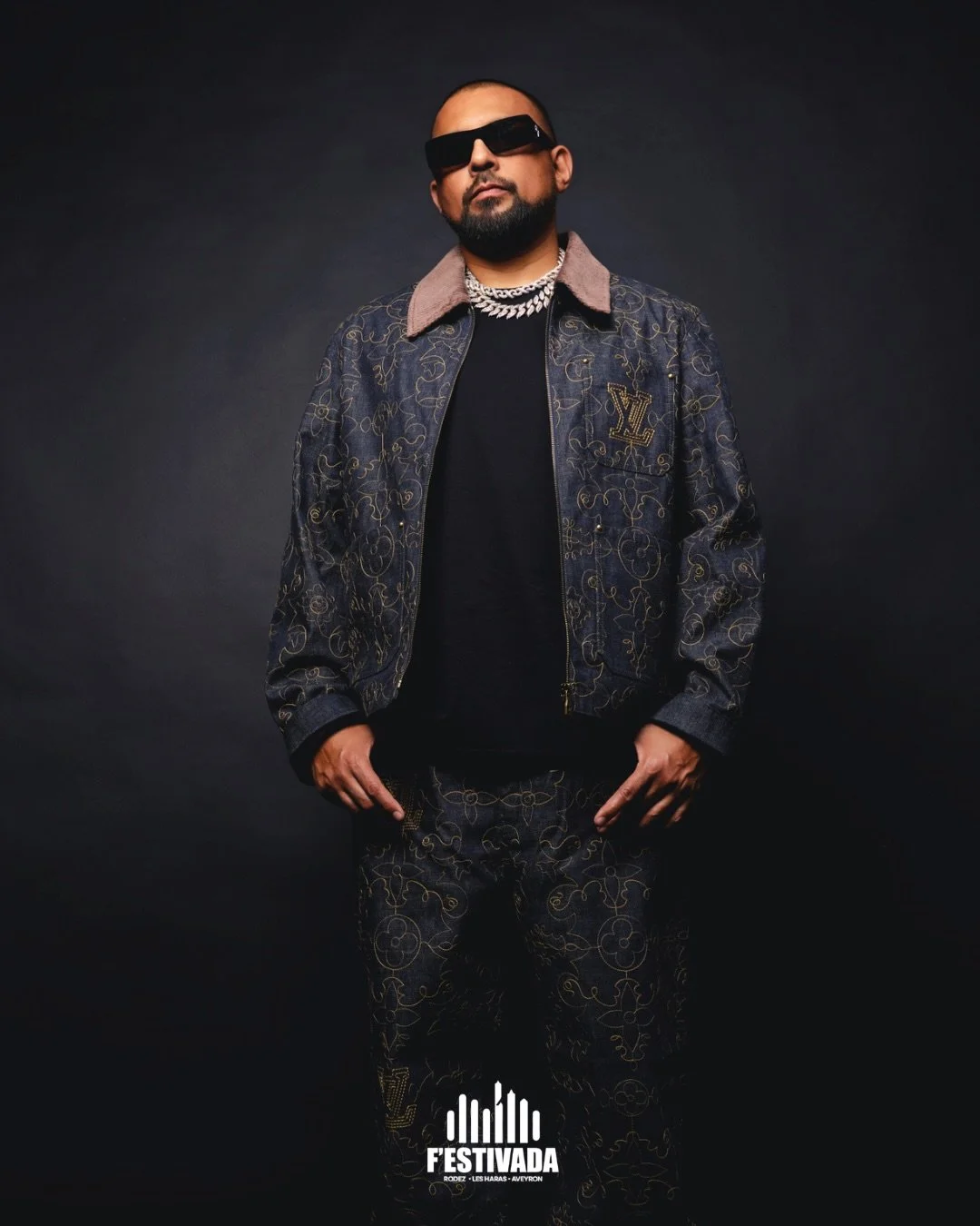 A man wearing black sunglasses, a designer Louis Vuitton denim jacket with gold embroidery, a black shirt, and matching patterned pants, posing against a dark background.