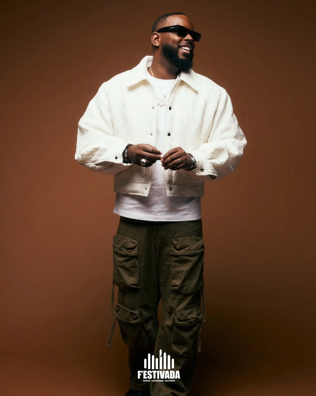 A smiling man wearing black sunglasses, a white jacket, a white t-shirt, and cargo pants, standing against a brown background with a logo that says 'FESTIVADA' at the bottom.