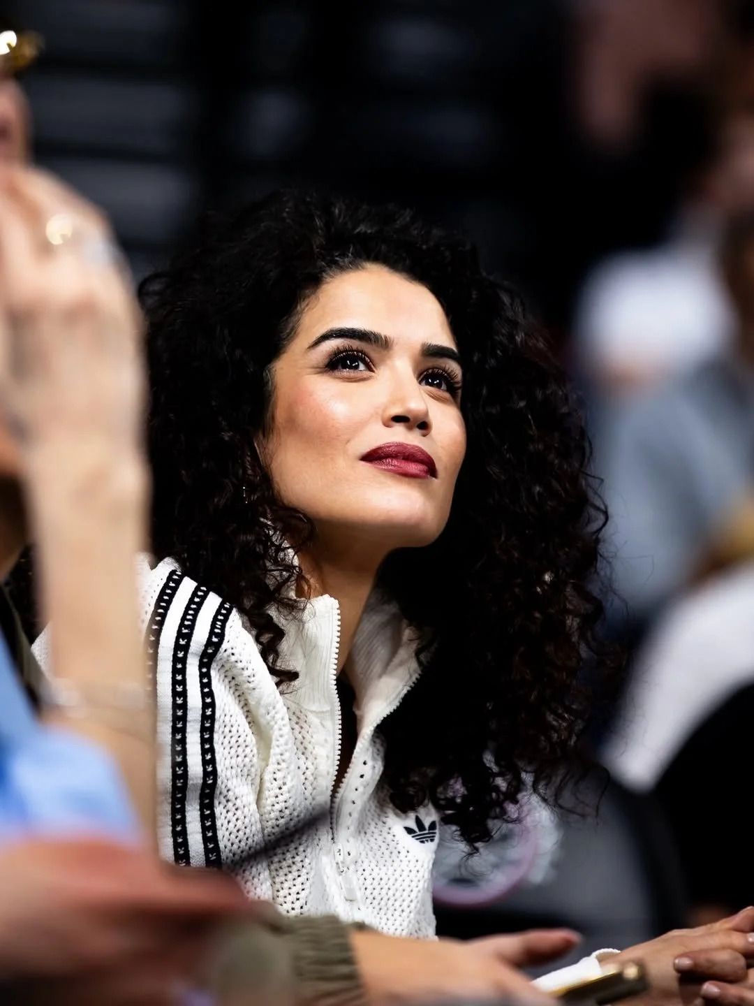 A woman with dark curly hair and wearing a white Adidas jacket with black stripes is looking upwards, sitting among other people in an indoor setting.