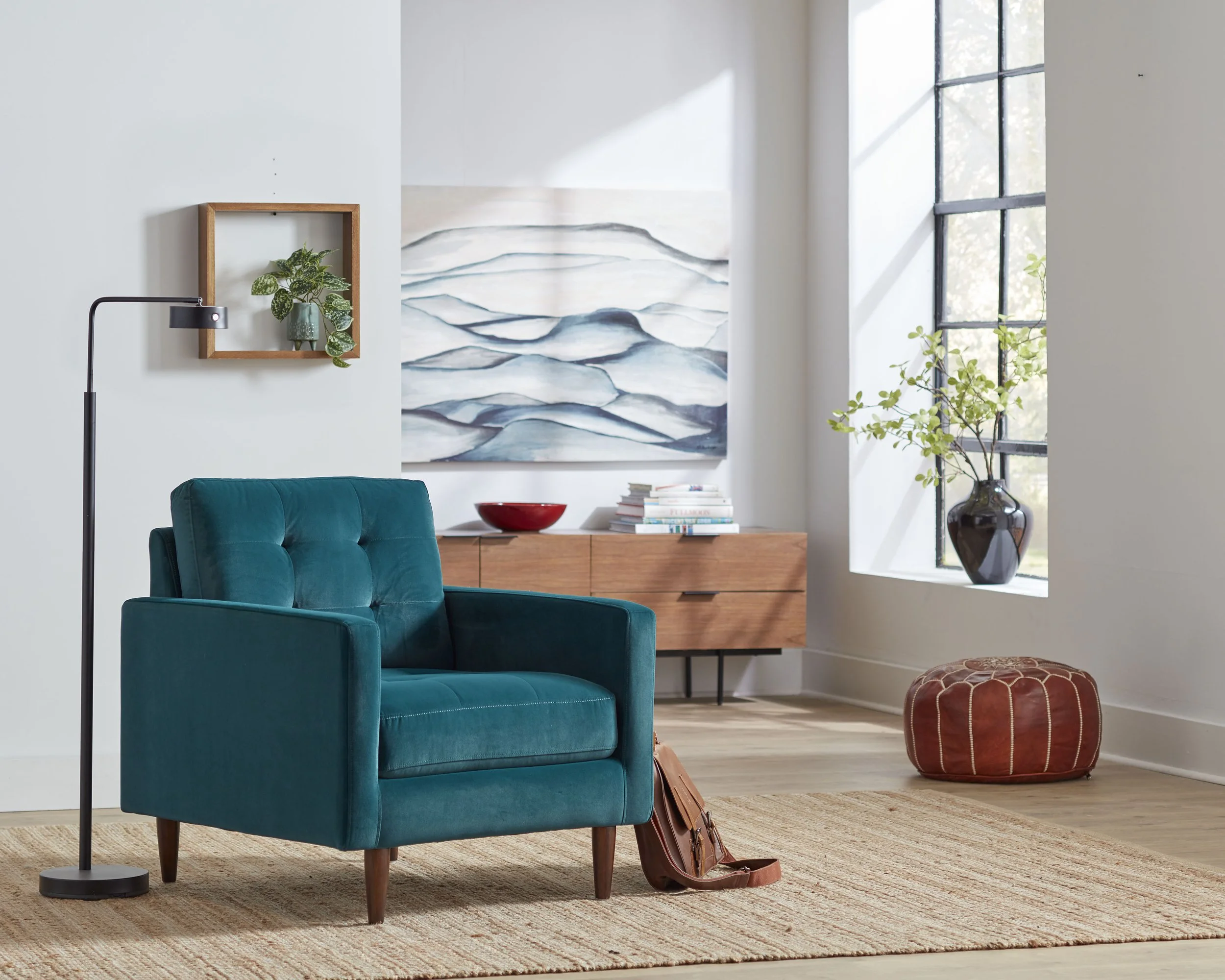 Living room with a teal armchair, a floor lamp, a wooden sideboard with books and a red bowl, a large window with sunlight, a potted plant in a black vase, and a painting of abstract blue and white waves on the wall.