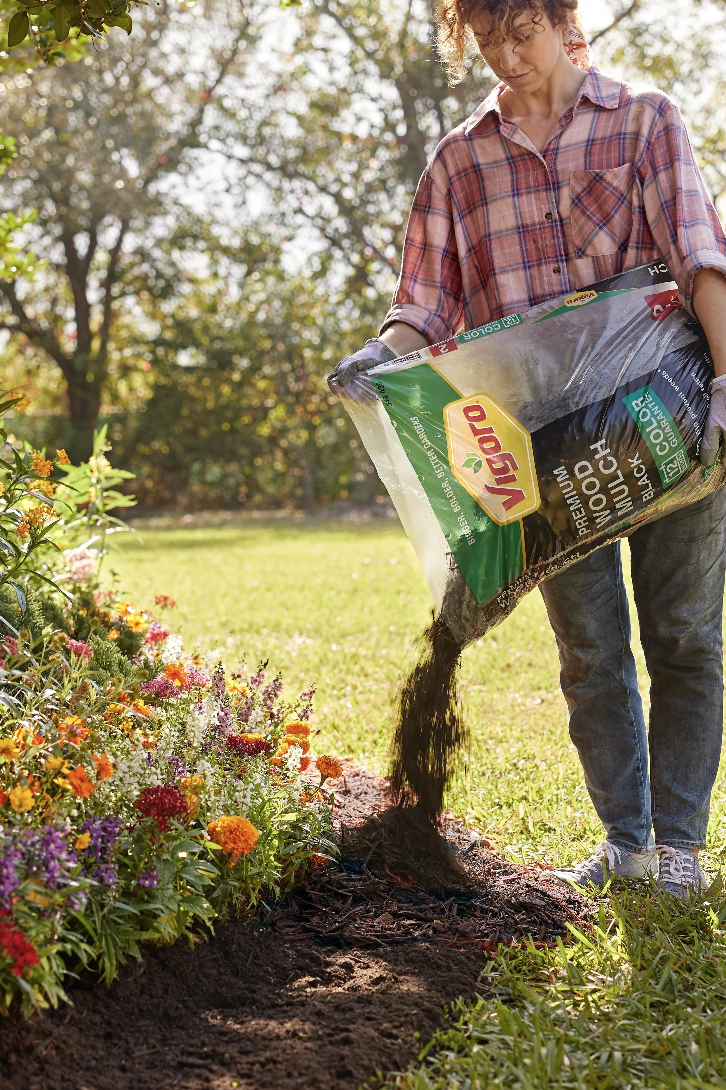 A woman in a plaid shirt and jeans is spreading mulch onto a garden bed with colorful flowers.