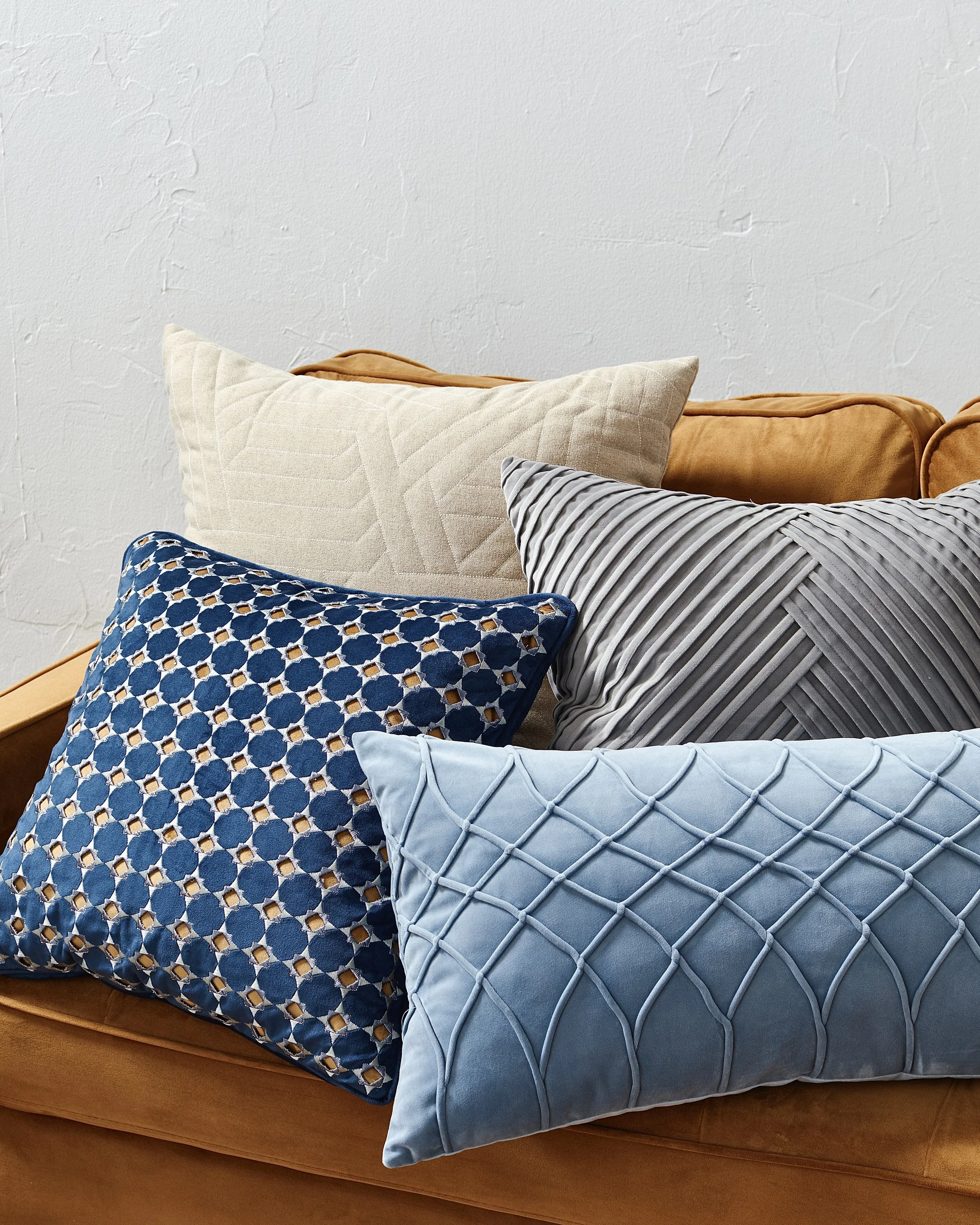 Five decorative pillows on a tan sofa, with a textured white wall in the background.